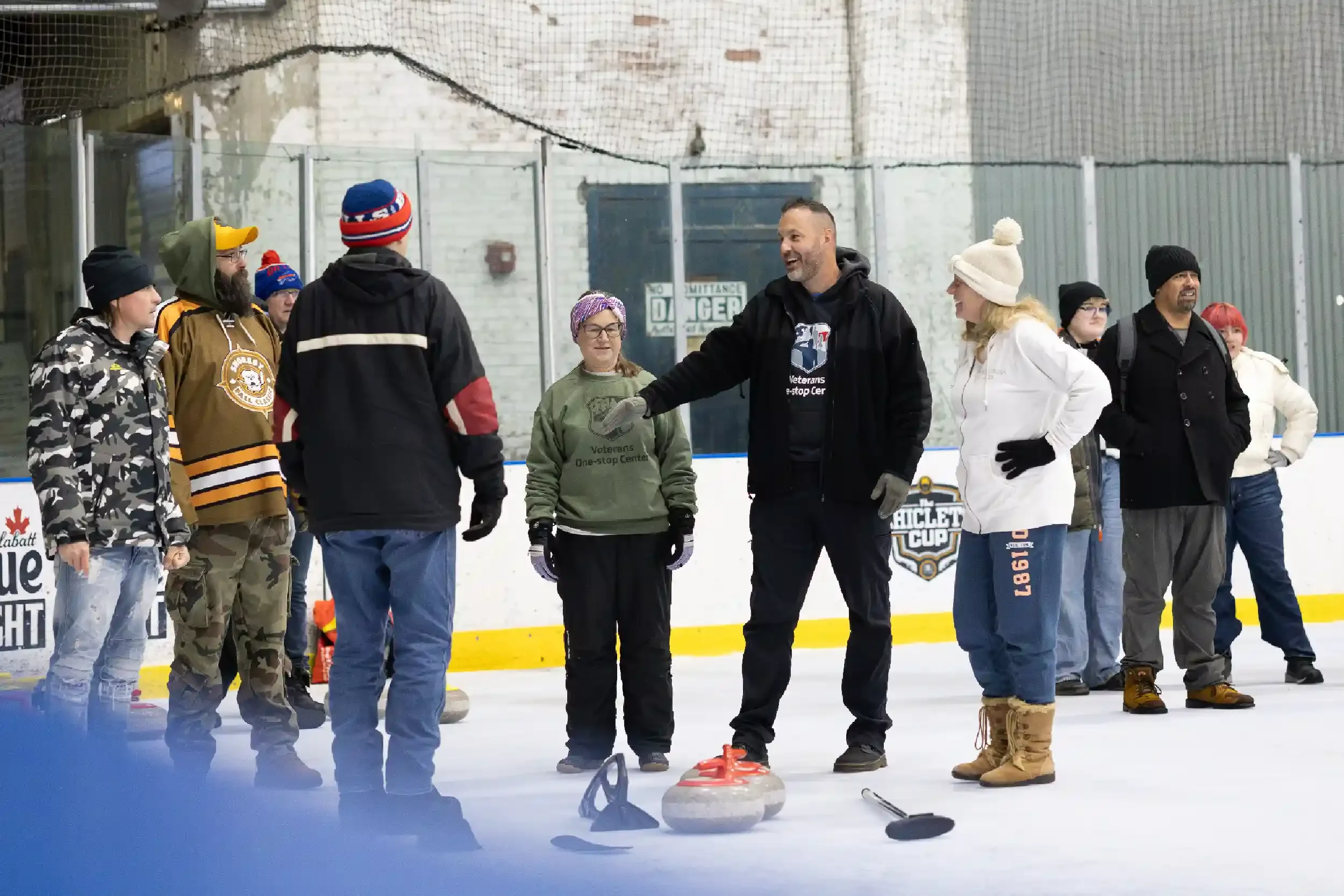 A group of people on an ice rink participating in a curling activity. A man in the center is explaining or instructing, surrounded by women and men dressed in winter clothing. Curling stones and a broom are visible on the ice.