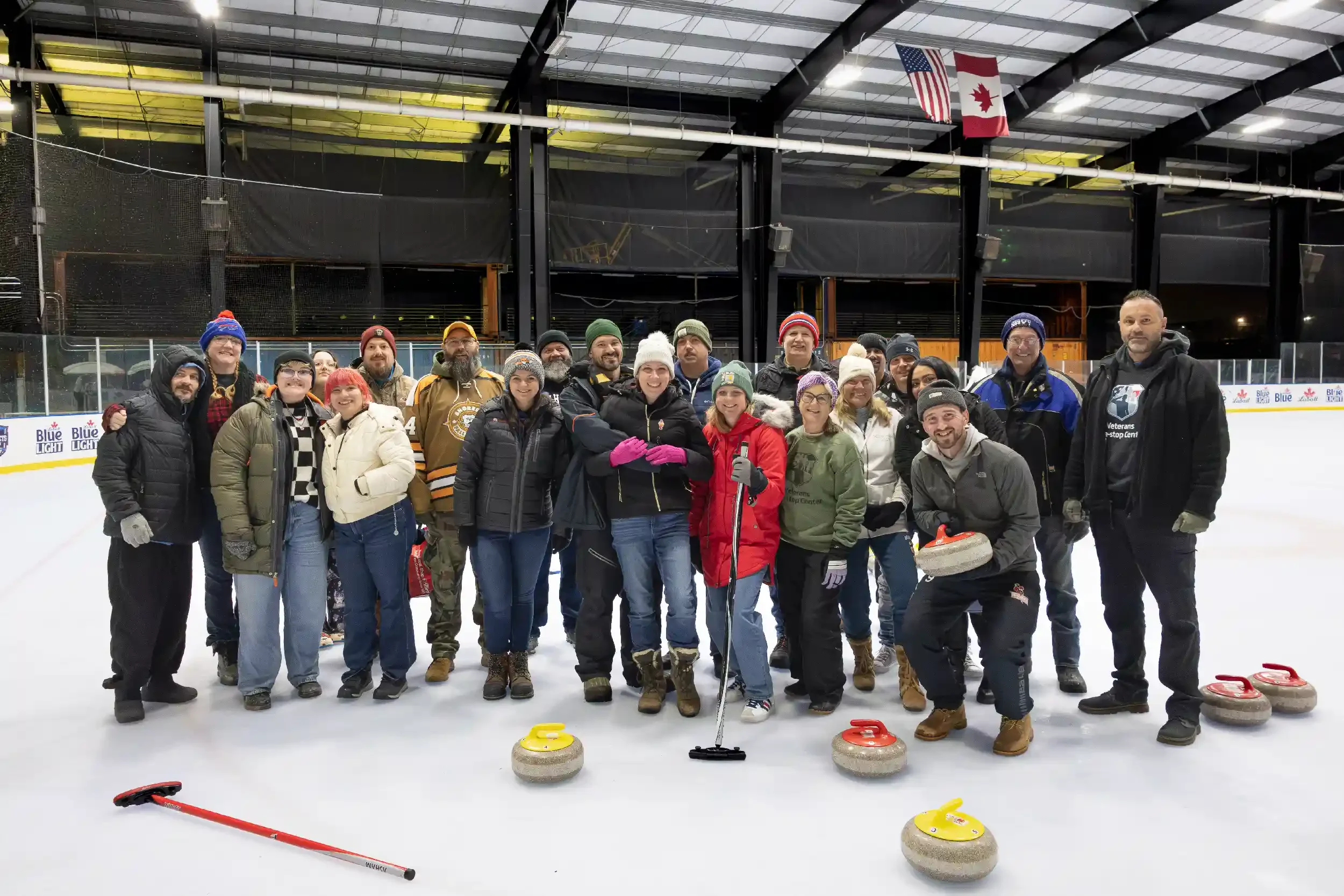 Group of people standing on an ice rink, wearing winter clothing, holding curling stones and a curling broom, with flags of the United States and Canada hanging from the ceiling.