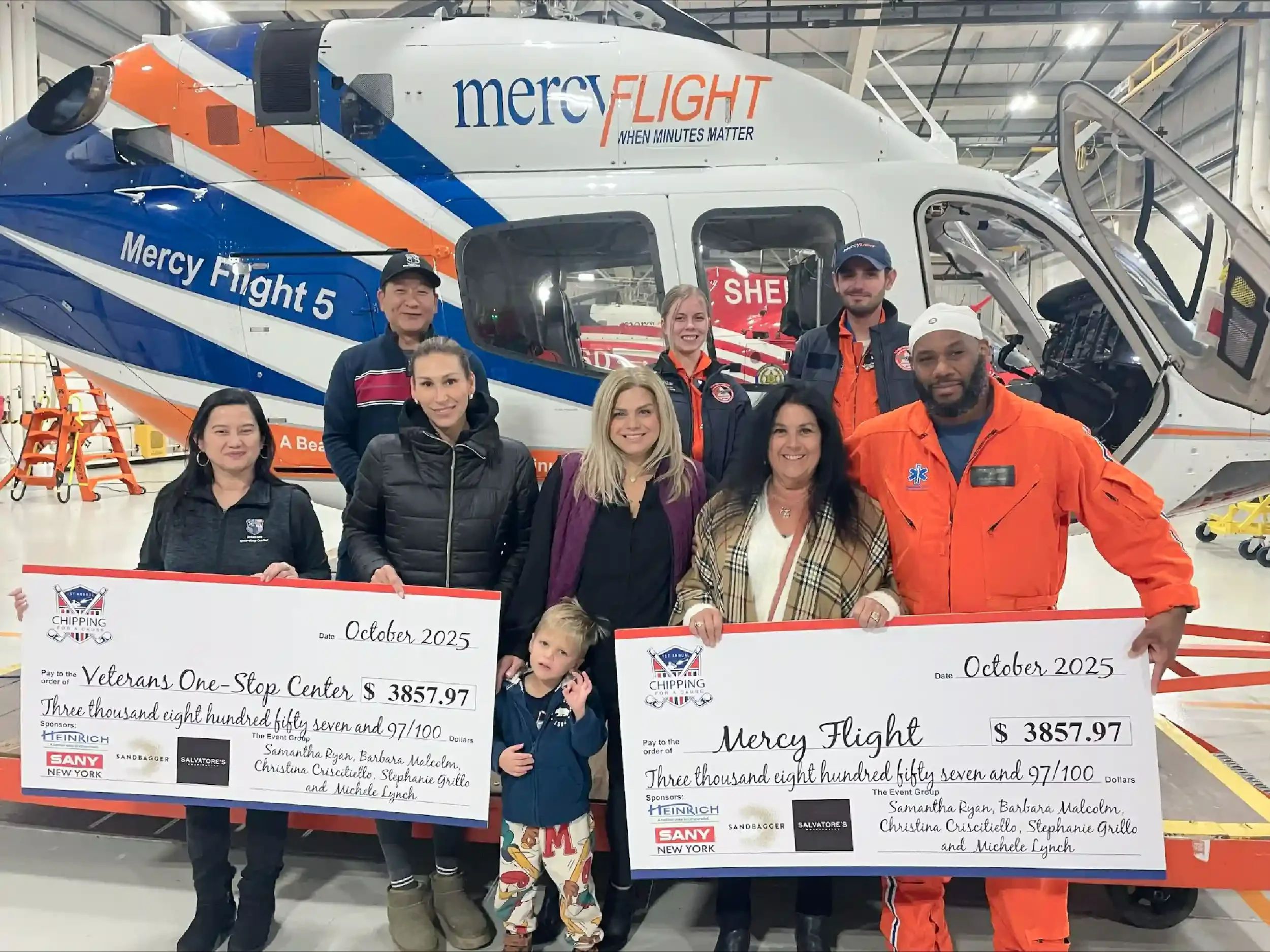 Group of people standing in front of a rescue helicopter holding oversized checks for $3857.97, with a young child in front. The helicopter has 'mercyFLIGHT' written on it, and the checks are payable to 'Veterans One-Stop Center' and 'Mercy Flight,' dated October 2025.