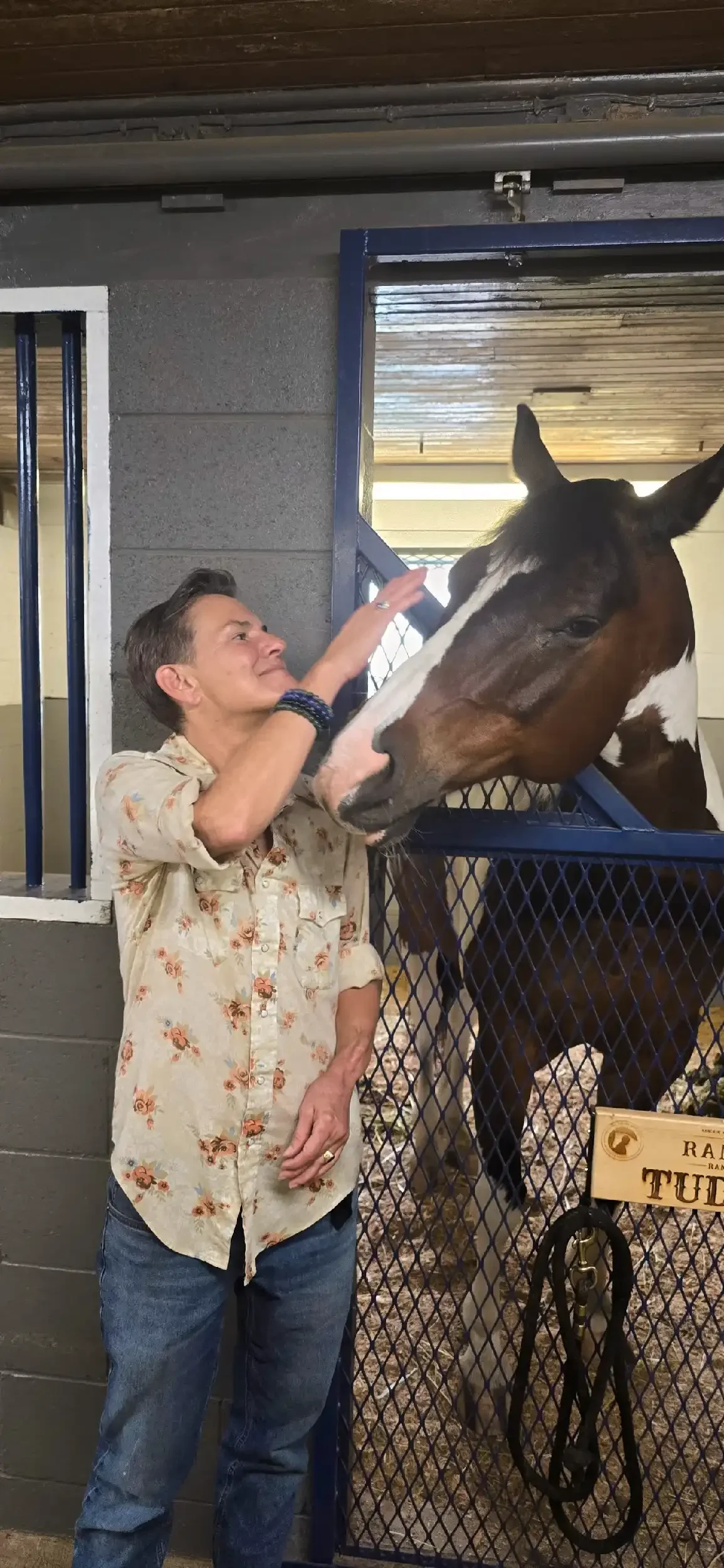 A woman with short hair wearing a cream floral shirt and jeans petting a brown and white horse inside a stable.