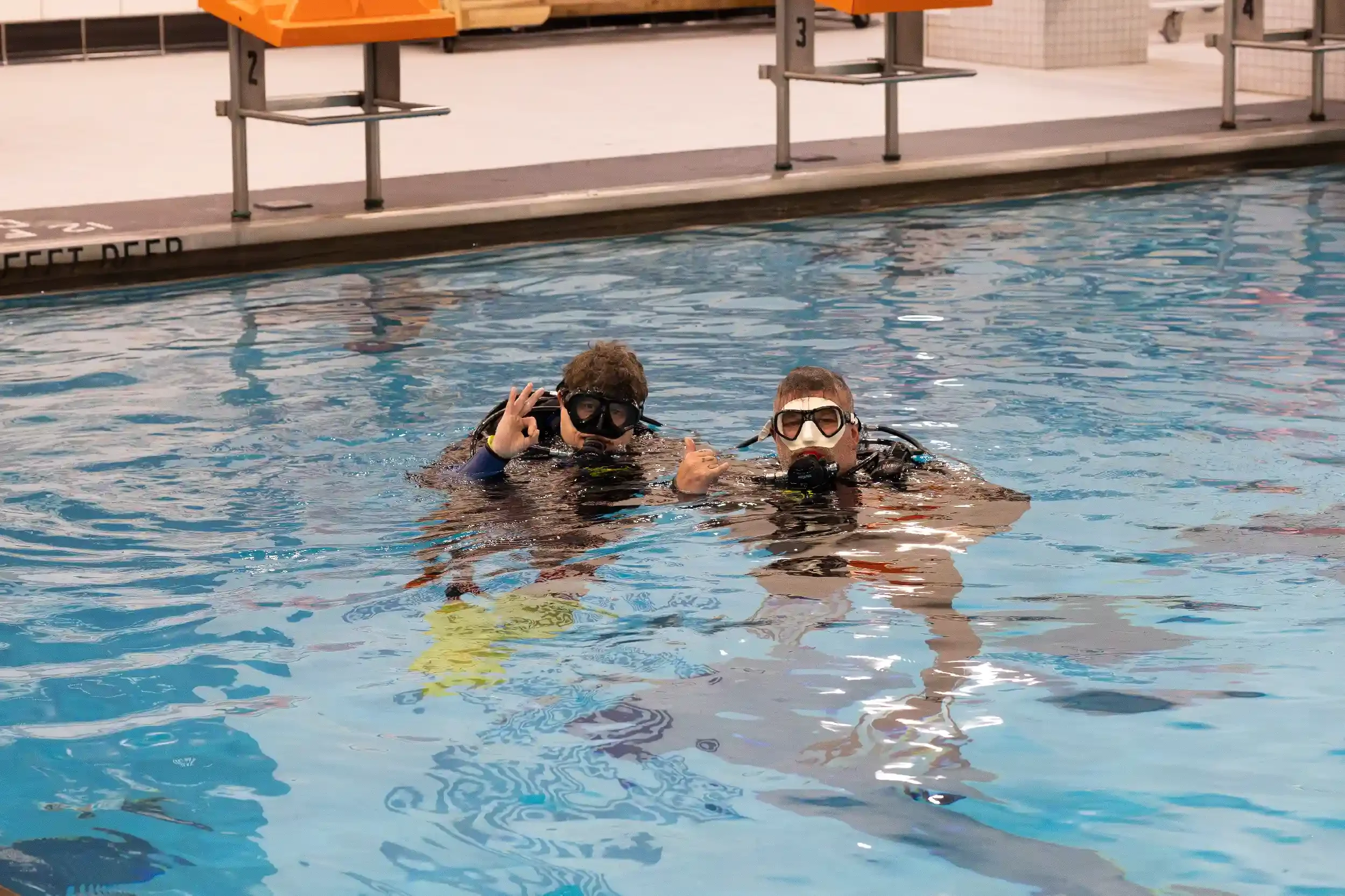 Two people in scuba gear floating in an indoor swimming pool, making the okay gesture and gesture of thumbs up.