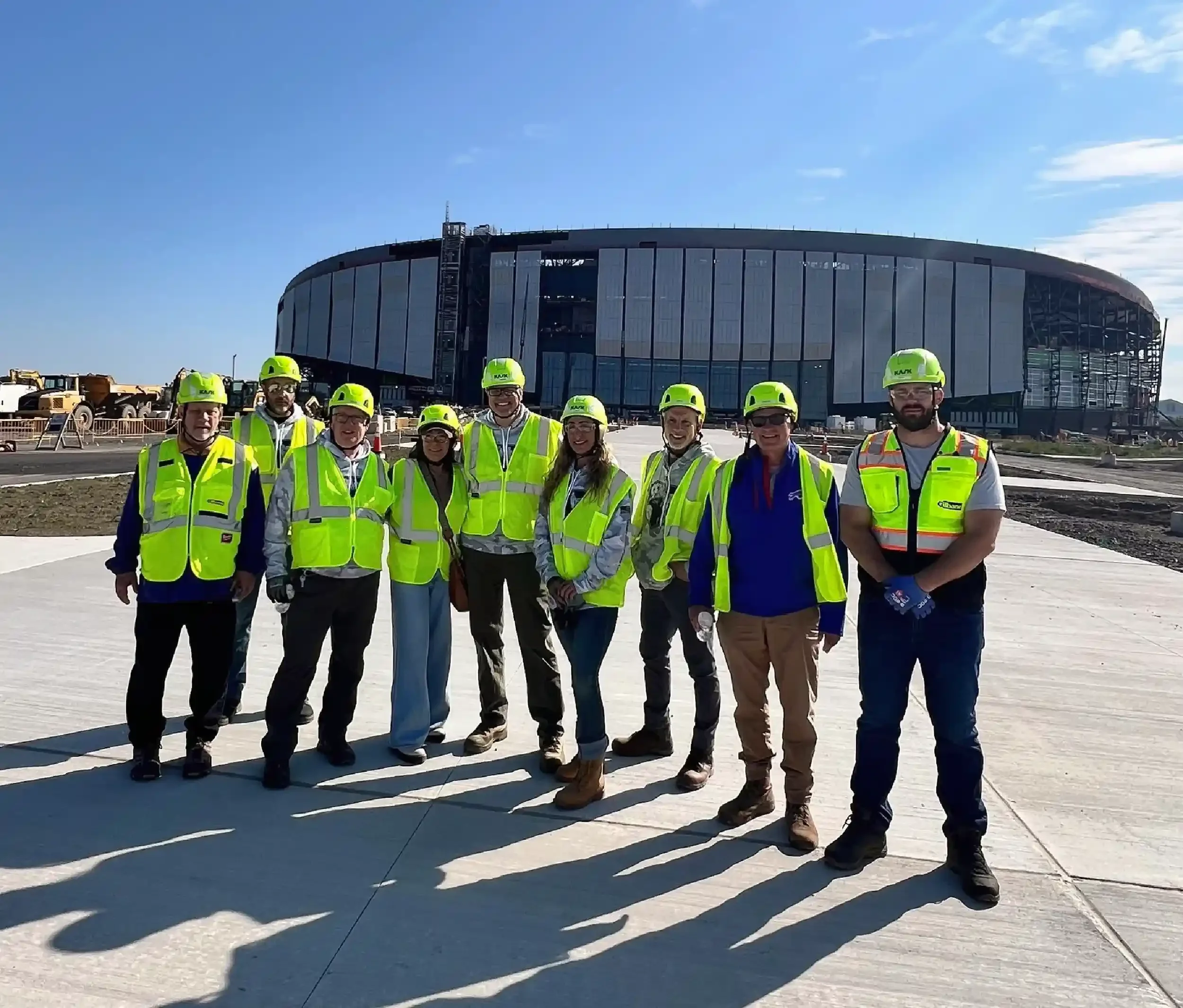 Group of construction workers and engineers wearing yellow safety vests and helmets standing on a construction site with a large stadium or arena building in the background under clear blue skies.