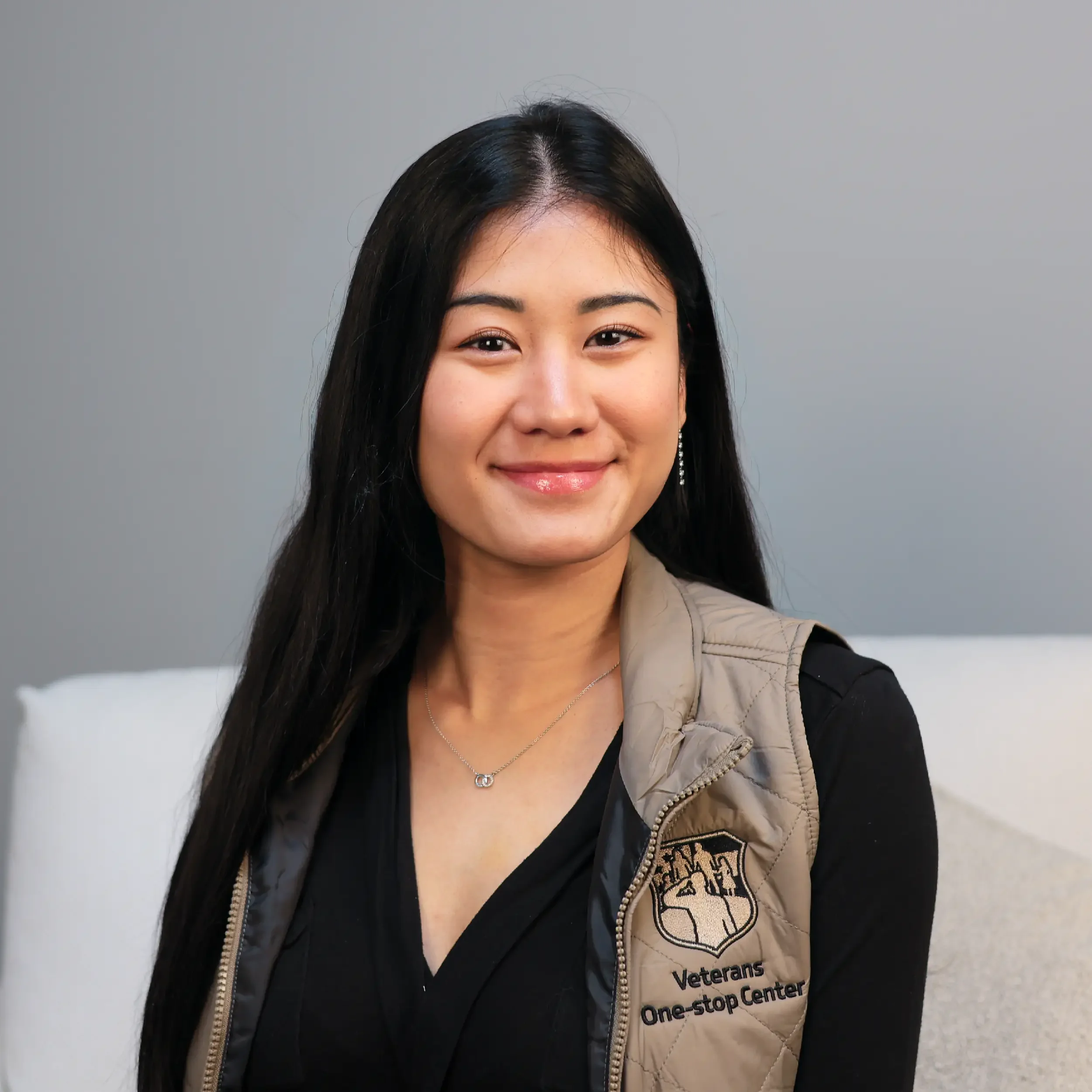 Portrait of a smiling woman with long black hair, wearing a beige vest with a 'Veterans One-Stop Center' logo and a black top against a gray background.
