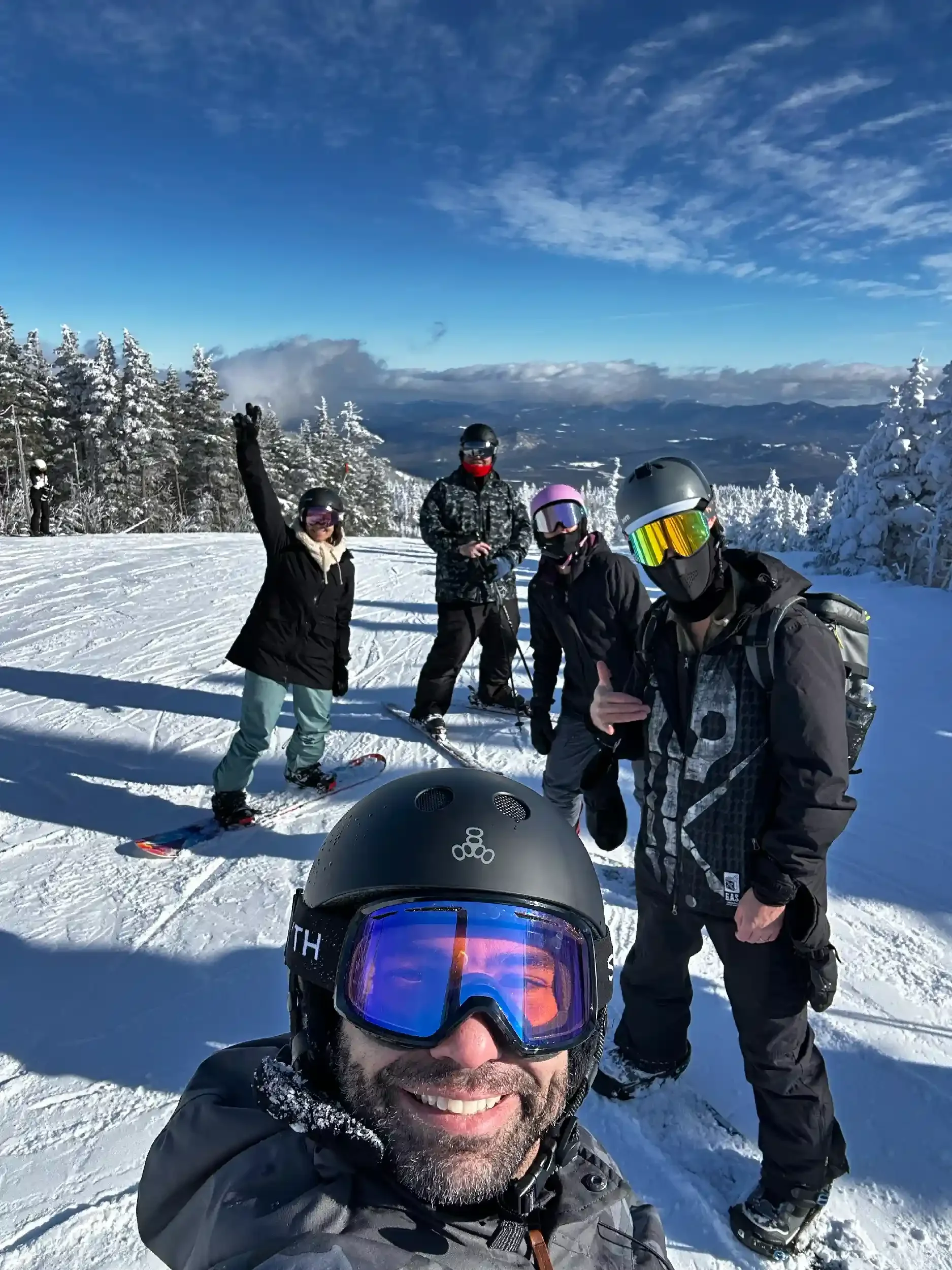 Group of five people skiing on a snowy mountain, taking a selfie, with snow-covered trees and a scenic mountain landscape in the background.