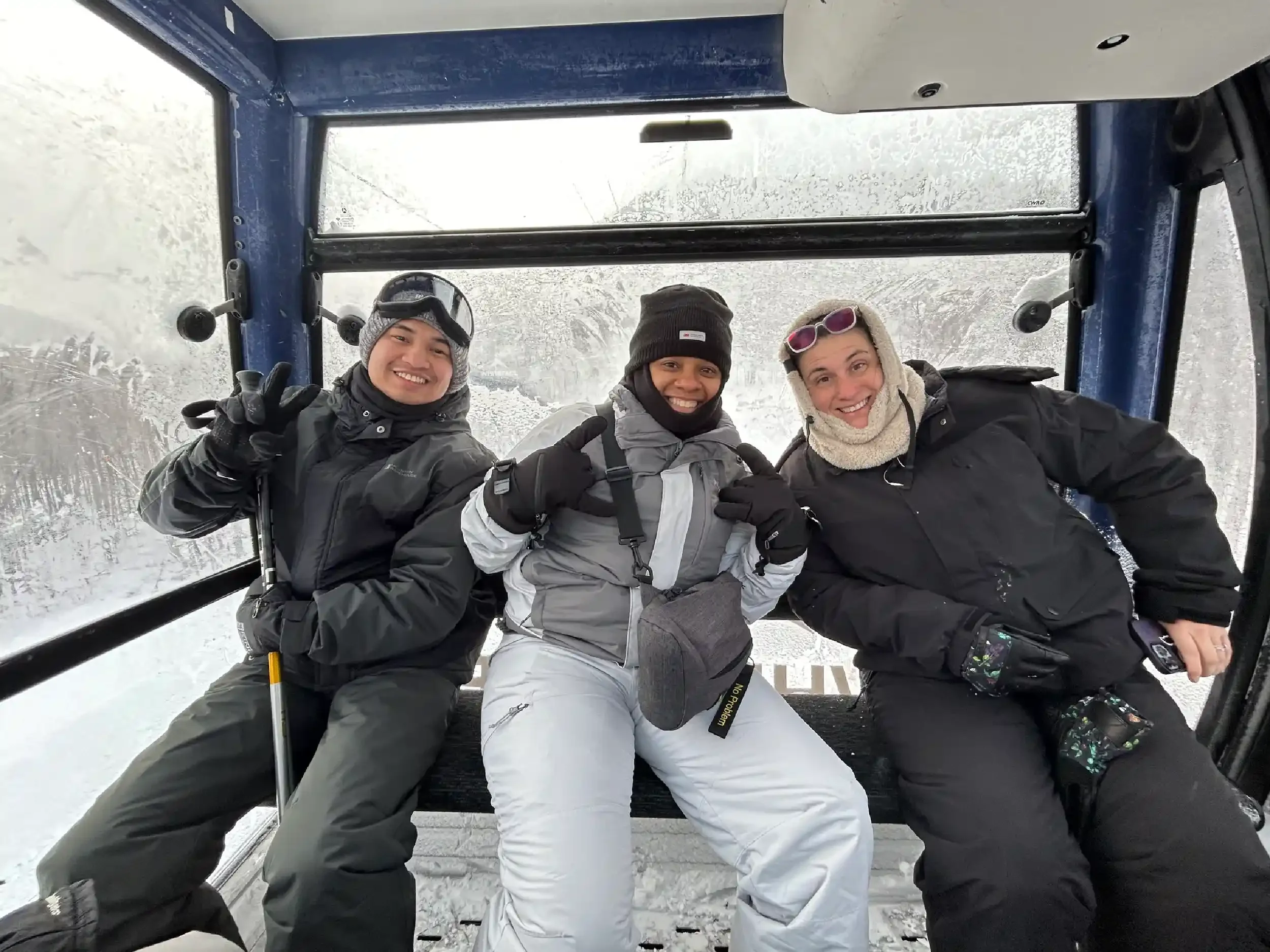 Three people sitting in a ski lift cabin with snowy background, smiling and making peace signs.