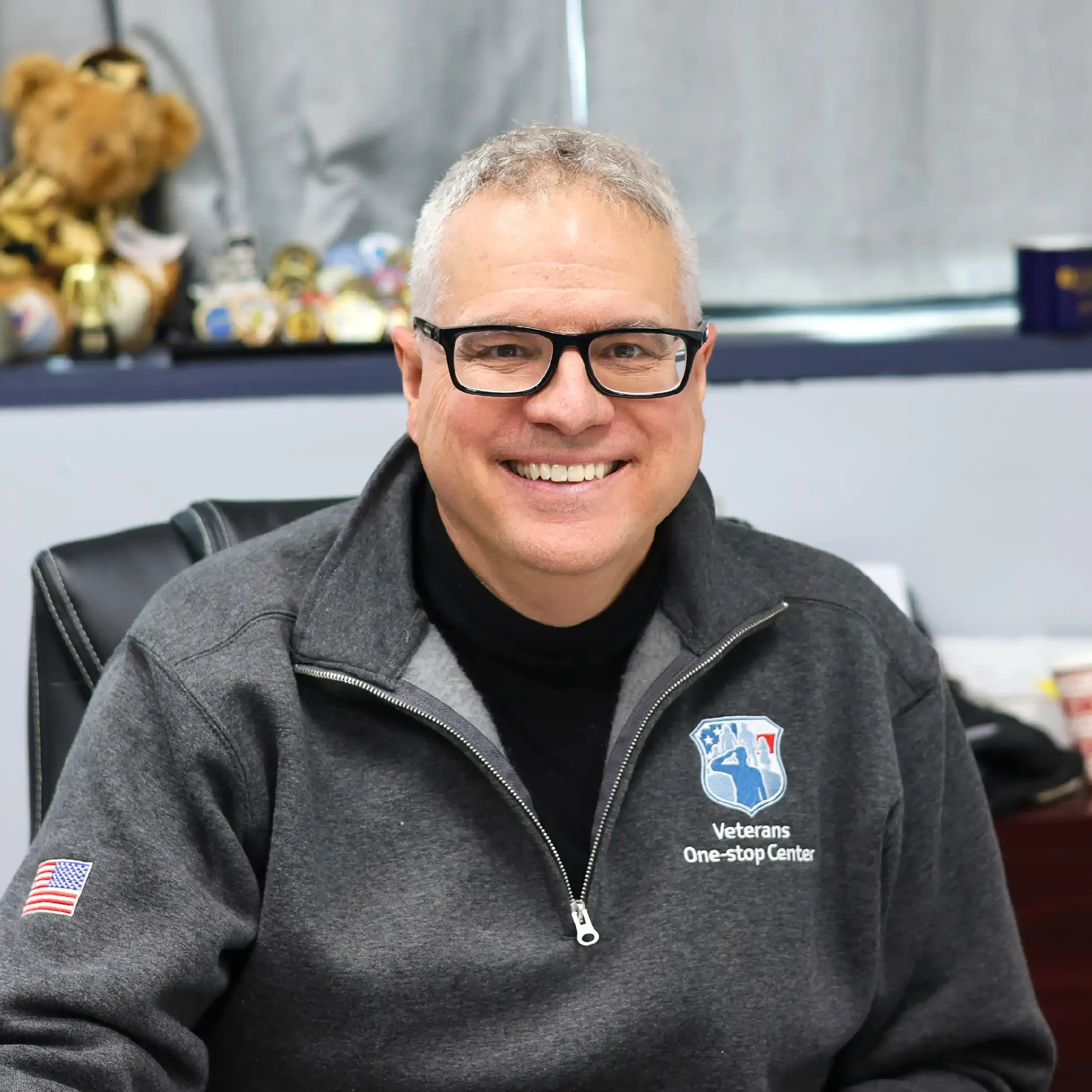 A smiling man wearing glasses and a dark gray jacket with a Veterans One-stop Center logo, sitting in an office with collectibles and teddy bears in the background.
