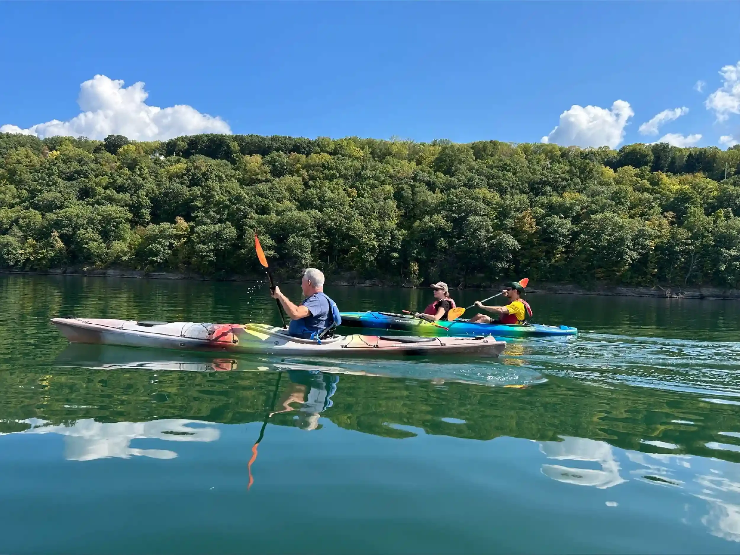 Three people kayaking on a calm lake with a forested hillside and a blue sky with clouds in the background.