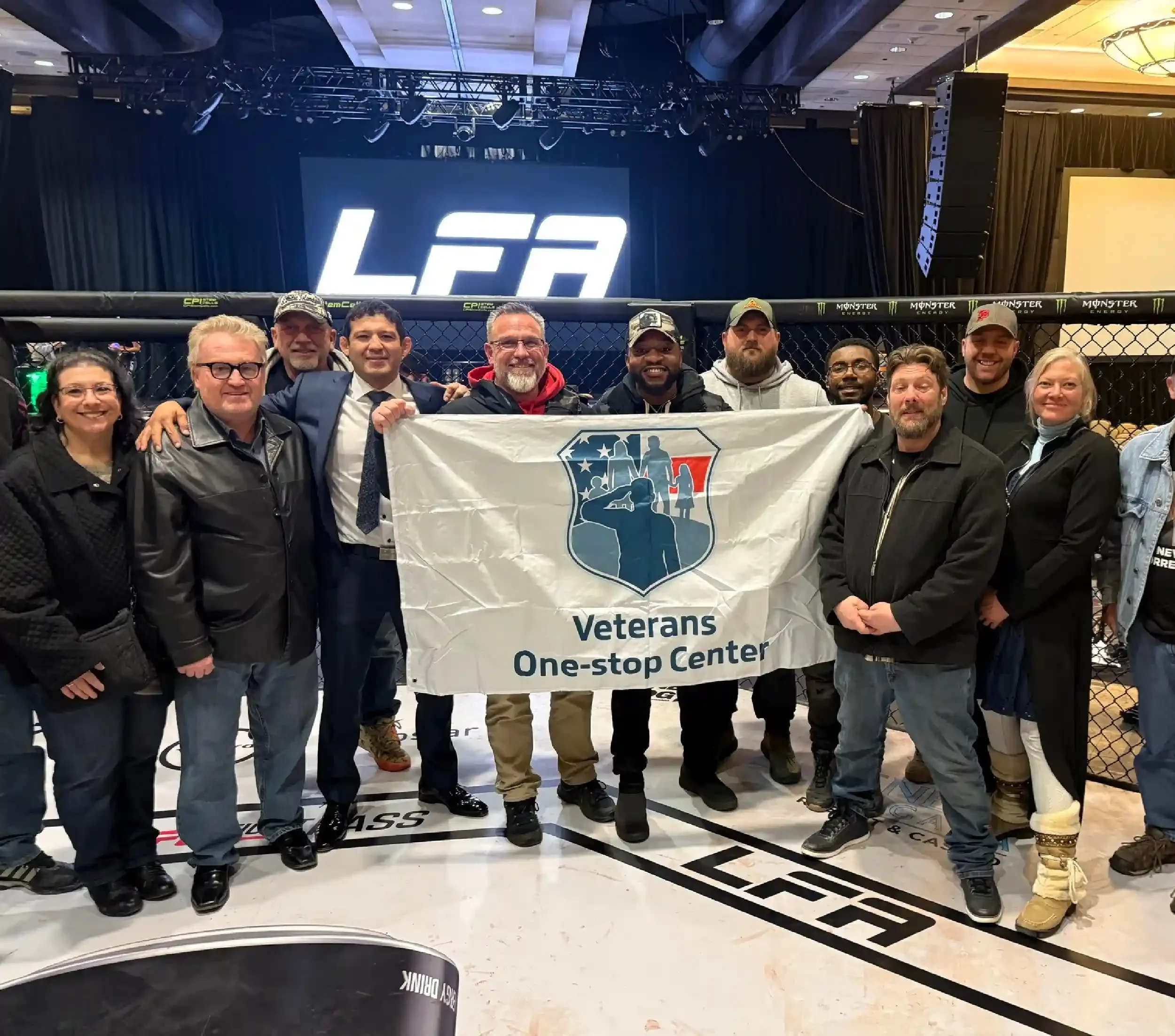Group of people standing inside a mixed martial arts cage holding a Veterans One-stop Center banner, with a large"LFA" logo on a screen behind them.