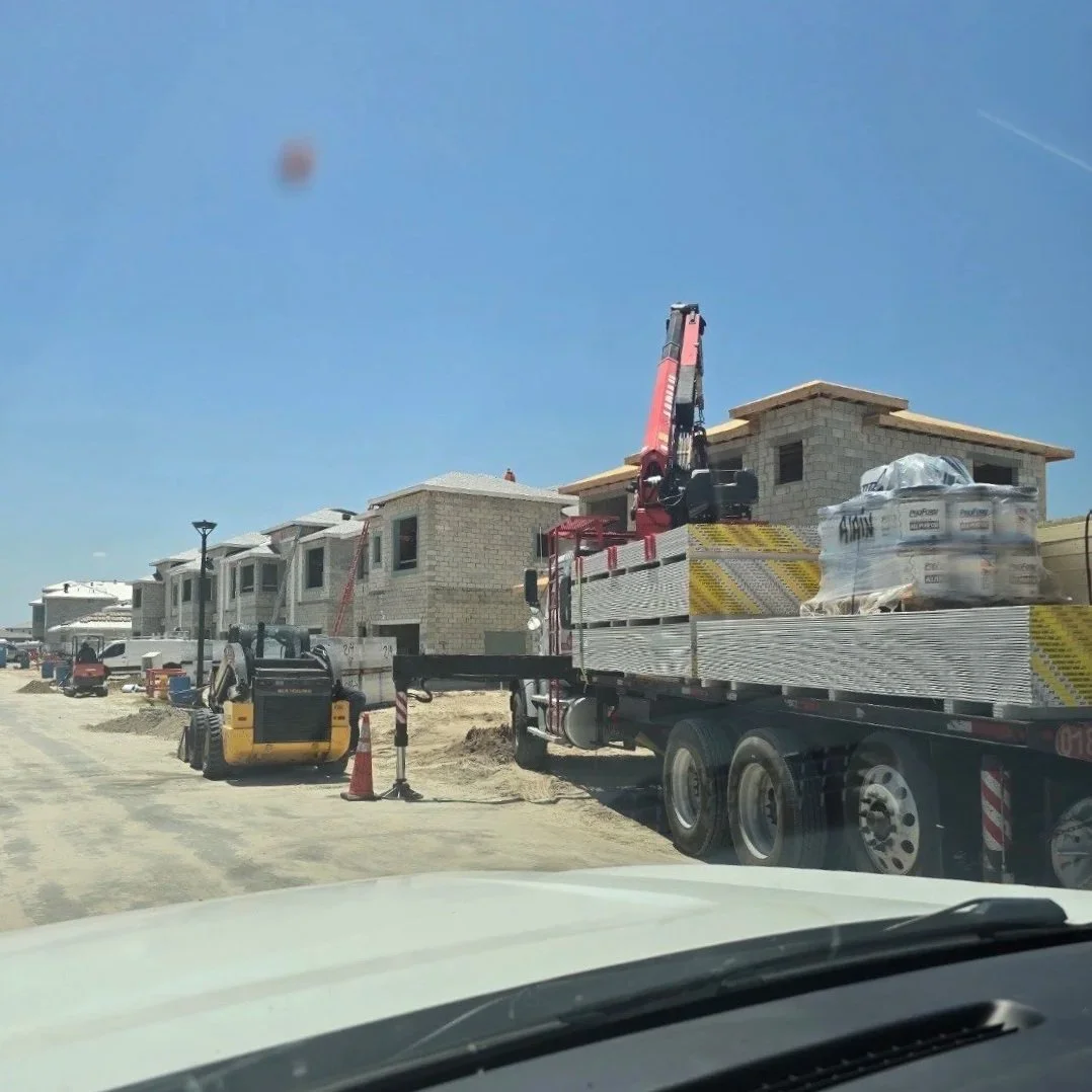 Construction site with unfinished houses, a flatbed truck with construction materials, a crane, and construction equipment under a clear blue sky.