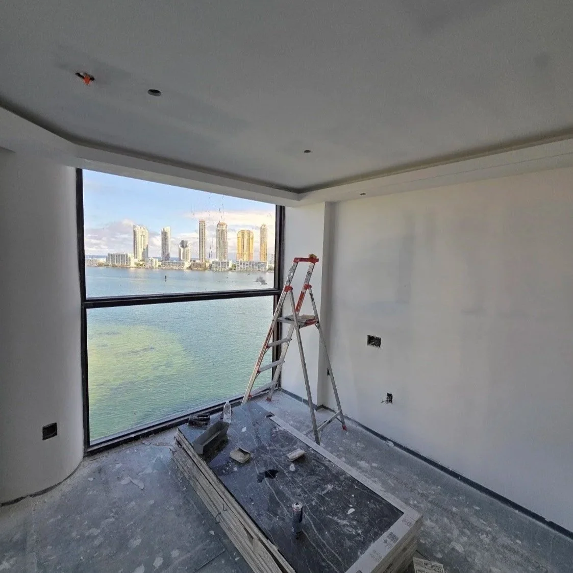 Interior of a high-rise apartment under construction with a large window overlooking a city skyline and water, a ladder, and construction materials on the floor.