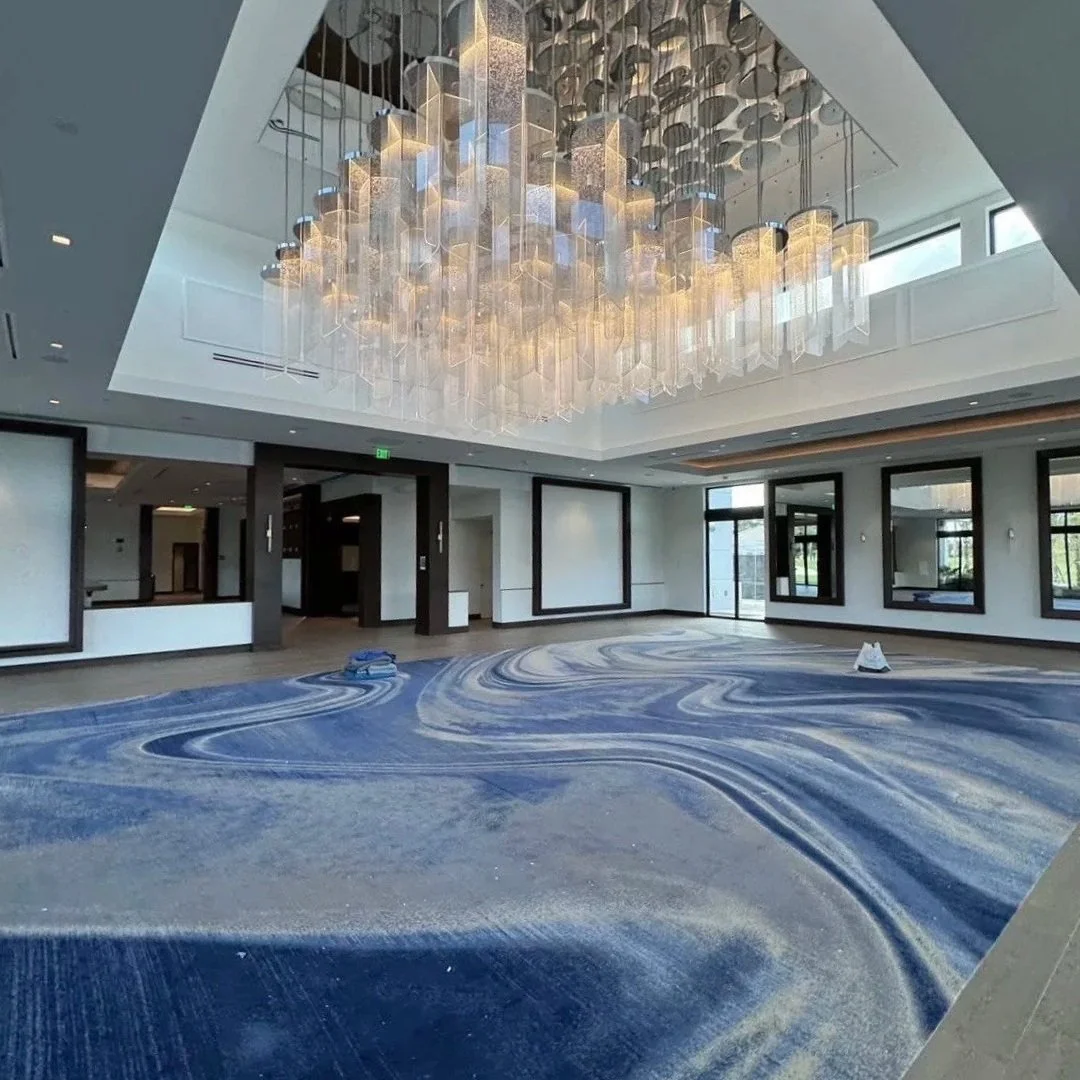 An empty modern conference or event room with a large blue and white swirling patterned carpet, a decorative ceiling with an elaborate chandelier, and large windows letting in natural light.