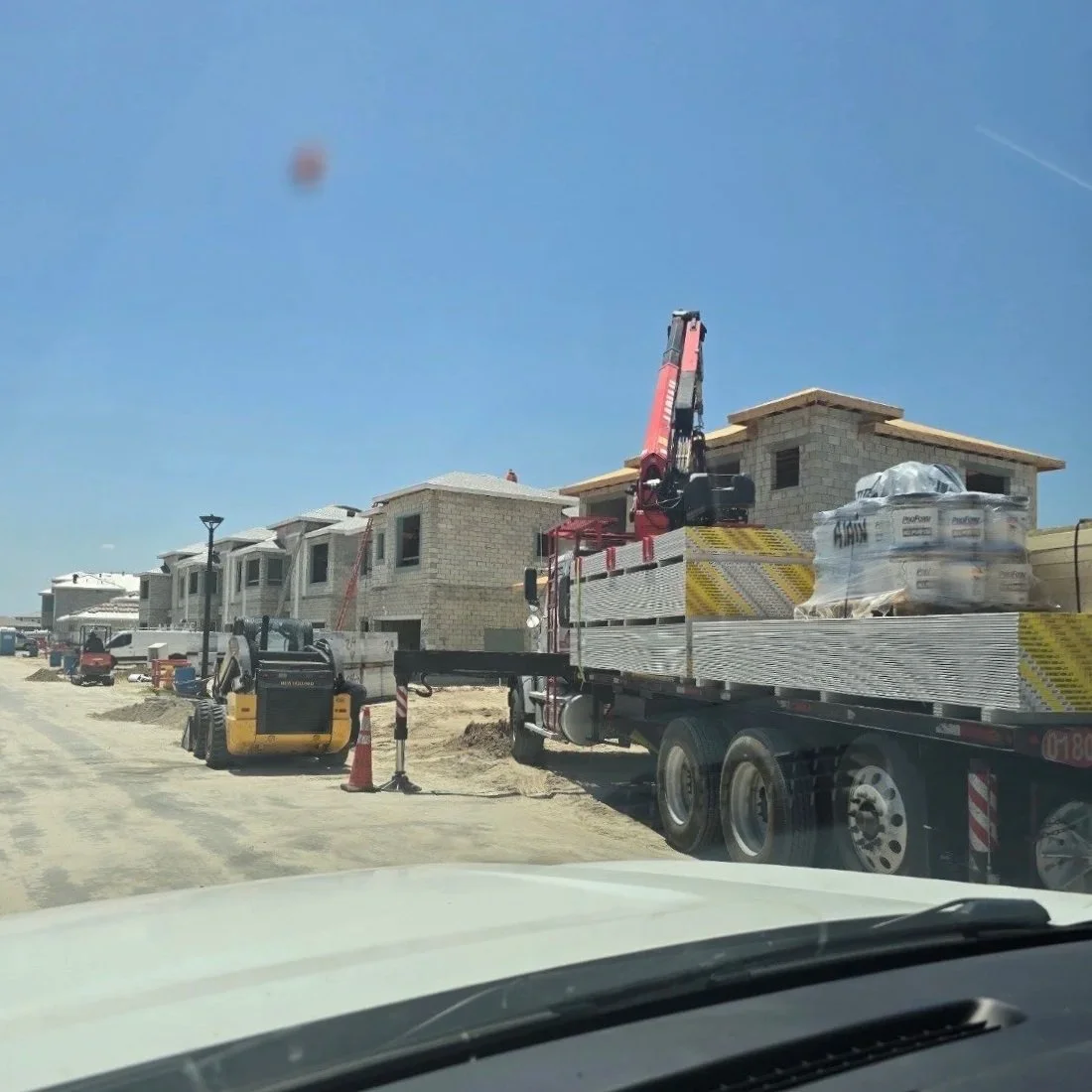 Construction site with partially built houses, a flatbed truck with construction materials, and construction equipment under a clear blue sky.