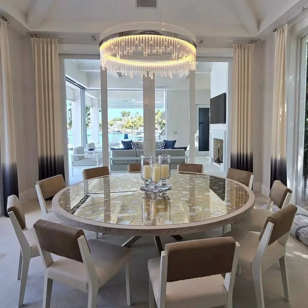 A modern dining room with a round table and ten beige chairs, a chandelier, and sliding glass doors leading to a patio and a view of water and palm trees outside.