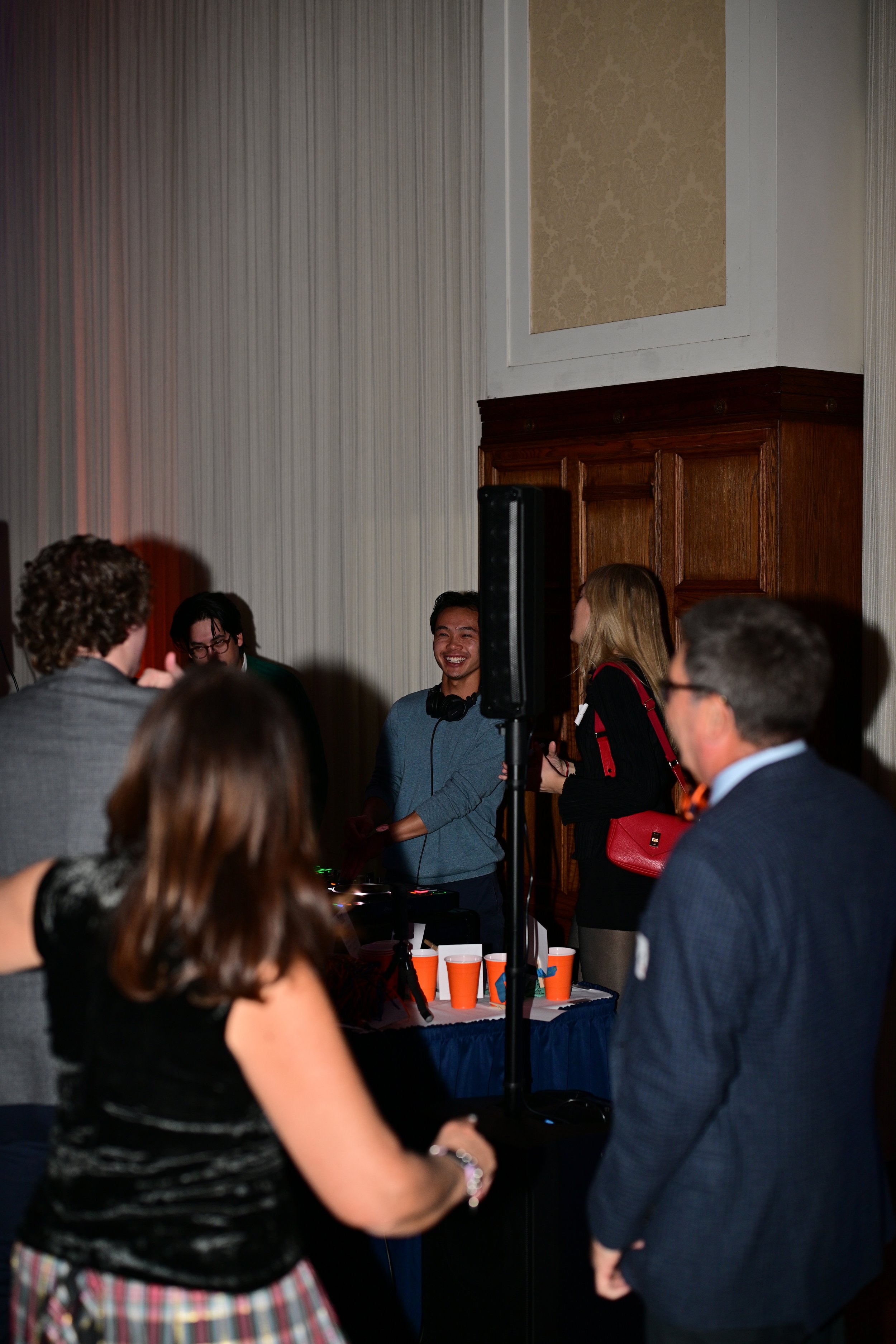People at a social gathering, some standing and others engaged in conversation, with a DJ setup and a speaker in the background.