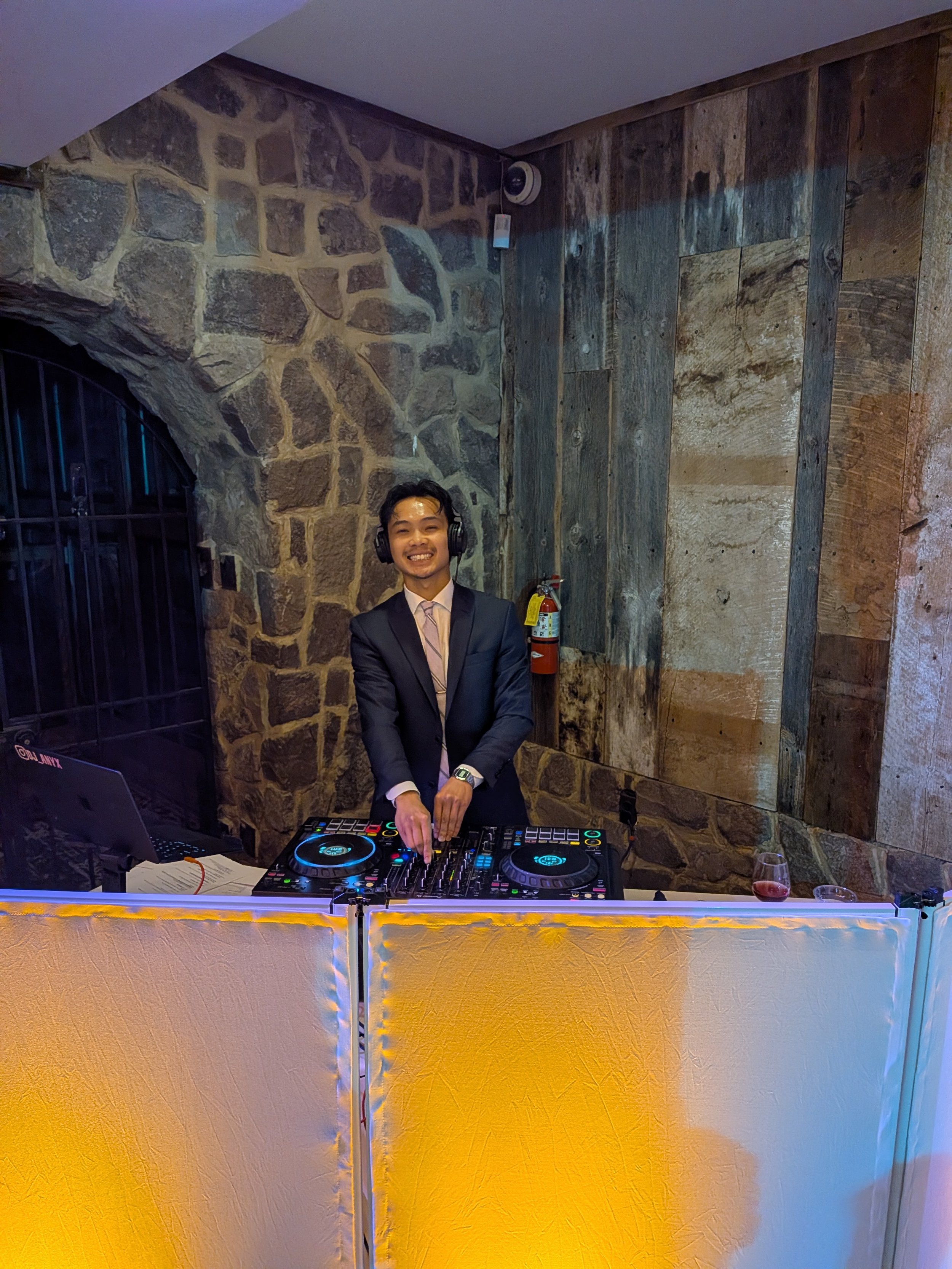 A man in a suit and tie smiling while DJing with turntables and a laptop at a party, with a stone wall and wooden panels in the background, and a glass of red wine on the bar.