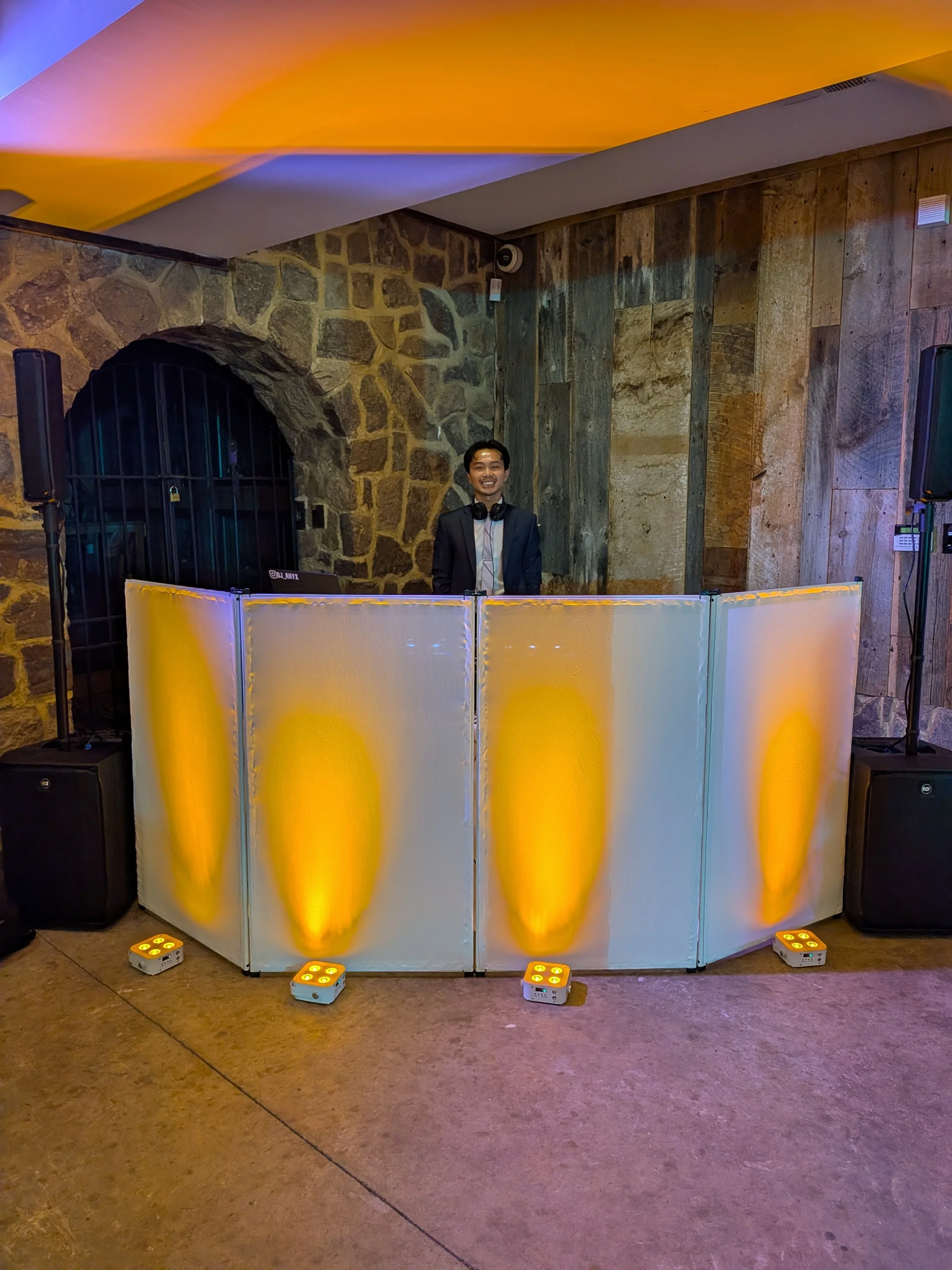 A man in a suit and headphones stands behind a DJ booth with yellow and white lighting effects in a rustic indoor venue with stone and wood walls.