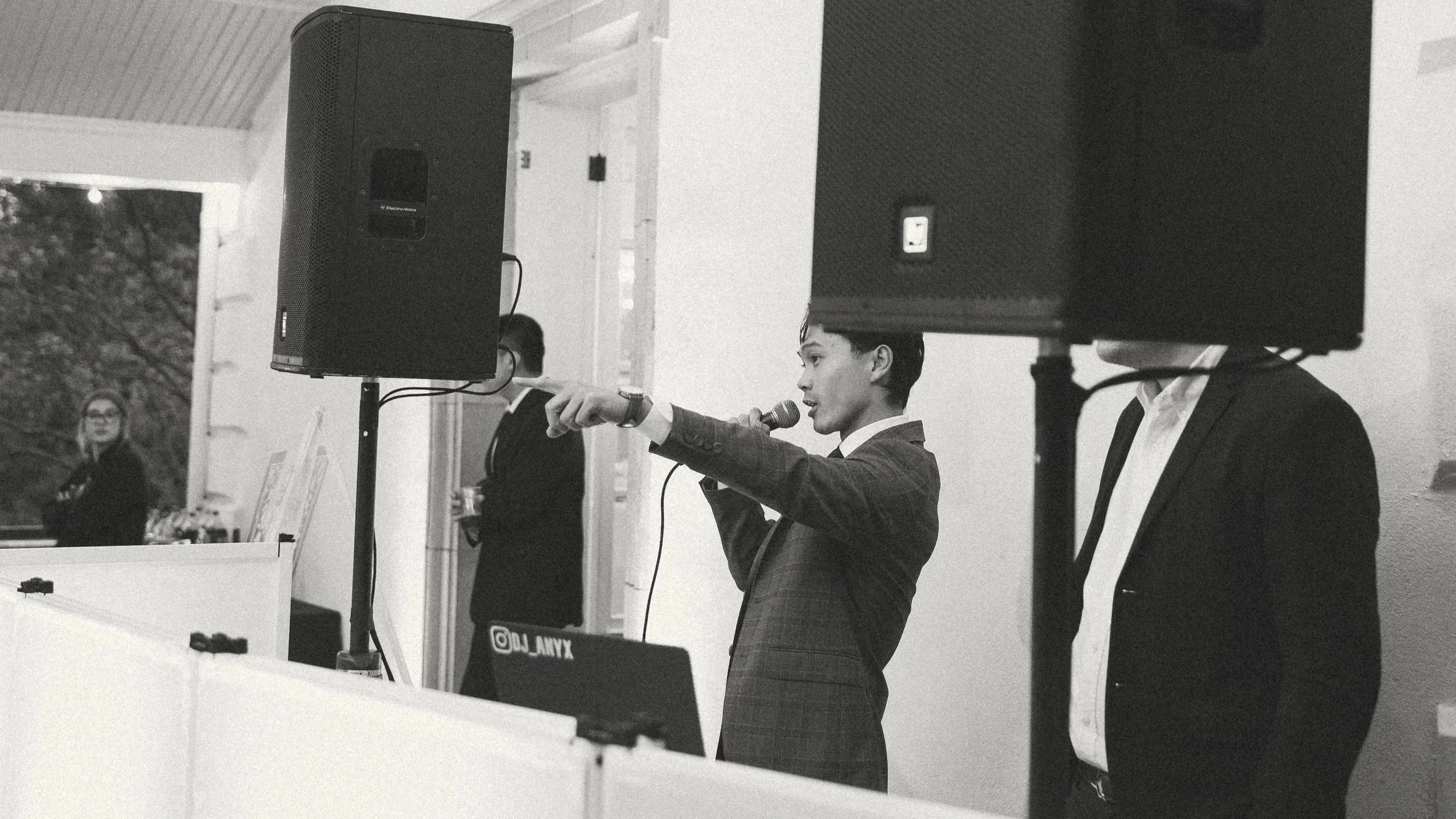 A young man dressed in a suit, speaking into a microphone and pointing with his finger at an event. He is standing behind speakers, with two women and another man in the background.