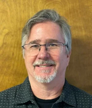 A middle-aged man with gray hair and glasses smiling, wearing a black shirt with white polka dots, standing against a wooden background.
