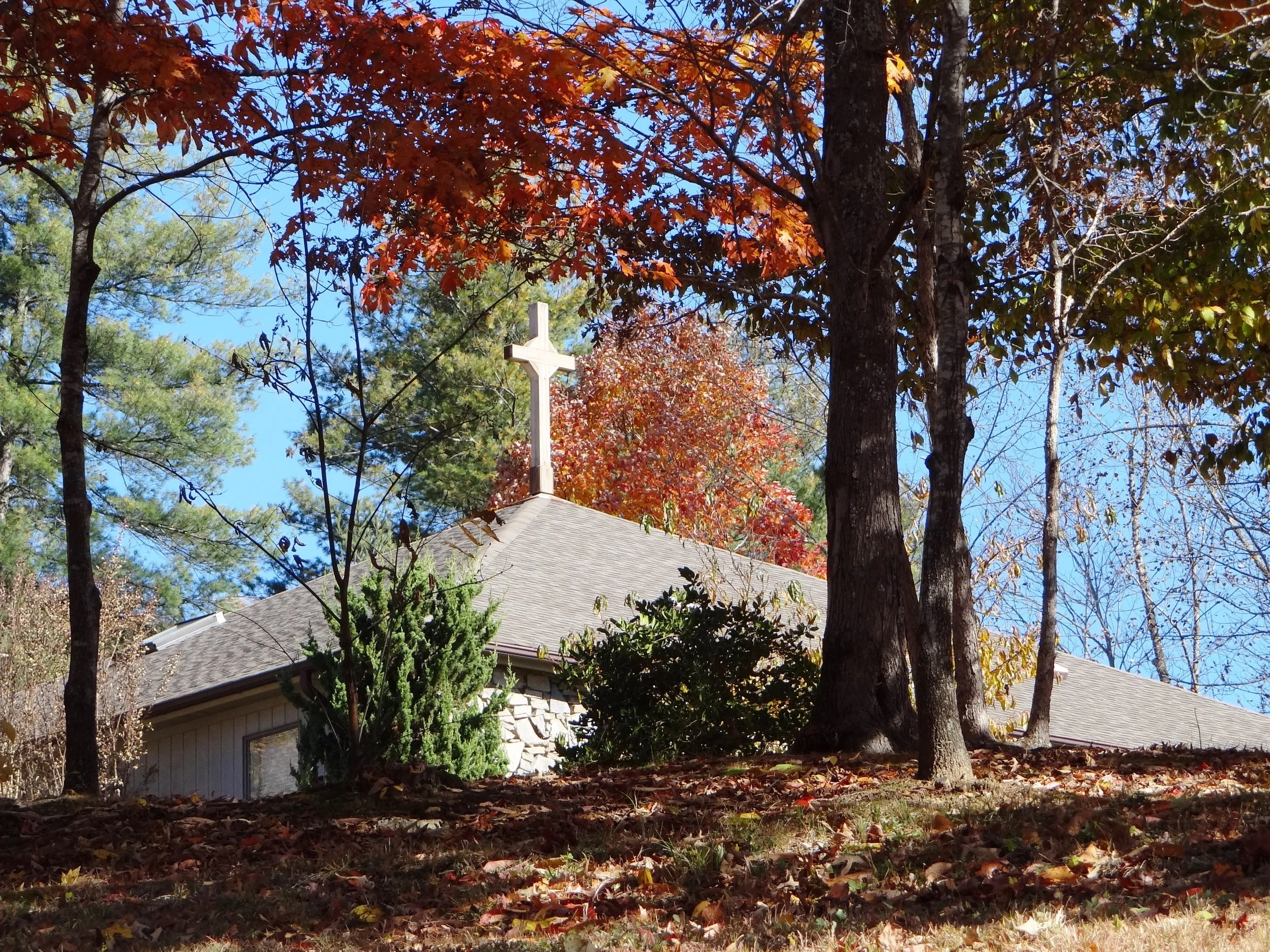 A church building with a white cross on the roof, surrounded by trees with fall foliage, in autumn.