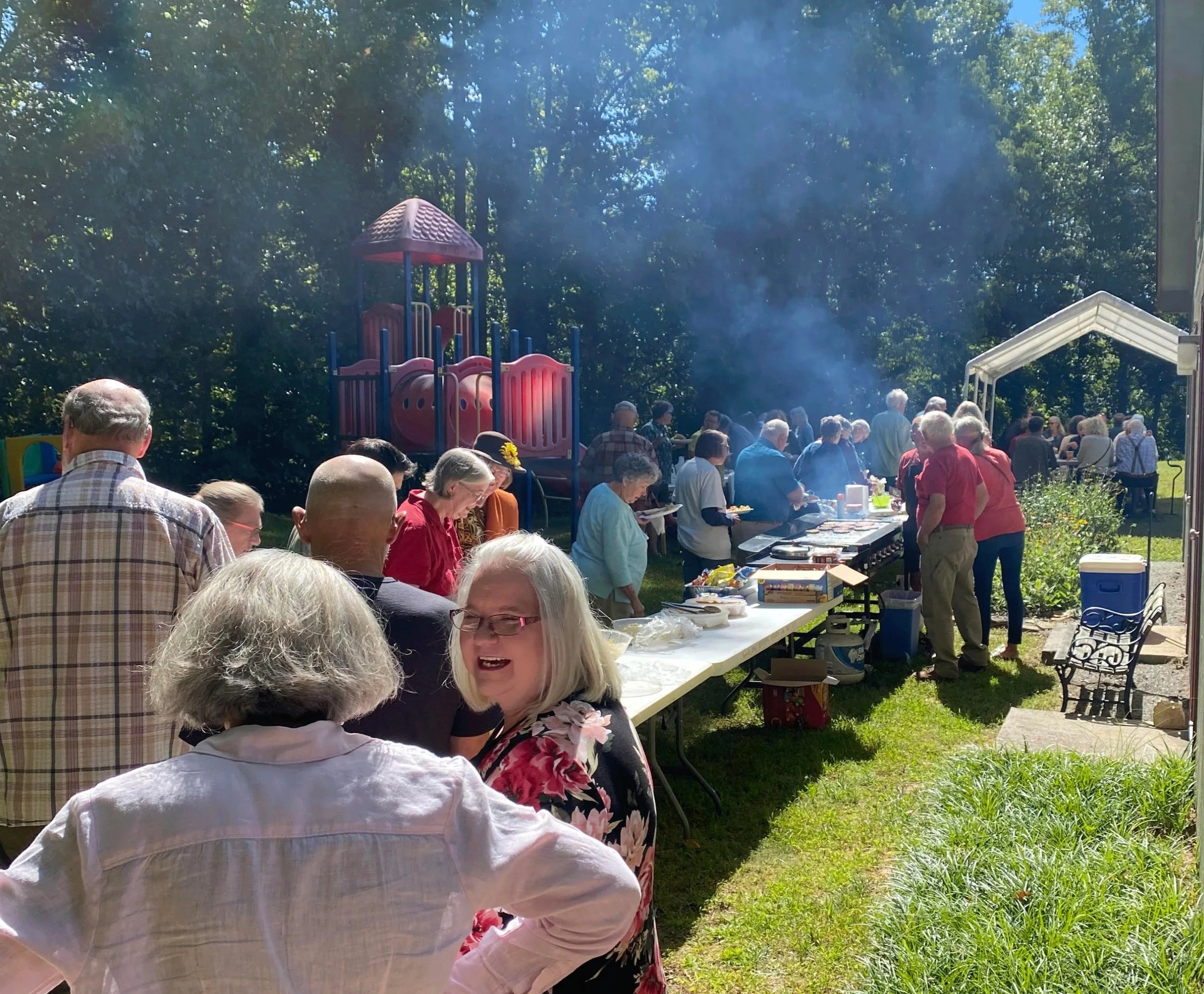 A large outdoor gathering with people around tables, a playground in the background, and a barbecue grill with smoke, on a sunny day.