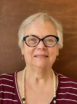 An older woman with short gray hair, glasses, wearing a striped top and a pearl necklace, standing in front of a wooden background.