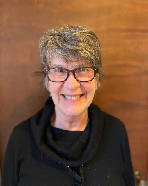 A smiling woman with short gray hair, wearing glasses and a black top, standing in front of a wooden wall.
