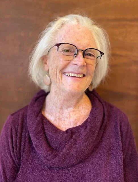An older woman with white hair, glasses, and pearl earrings, smiling in front of a wooden background, wearing a purple sweater.