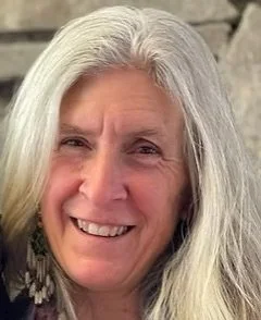 Close-up of a smiling woman with long white hair outdoors.