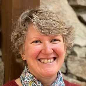 A smiling woman with short, curly gray hair, wearing a colorful patterned scarf and a maroon sweater, standing in front of a stone wall.
