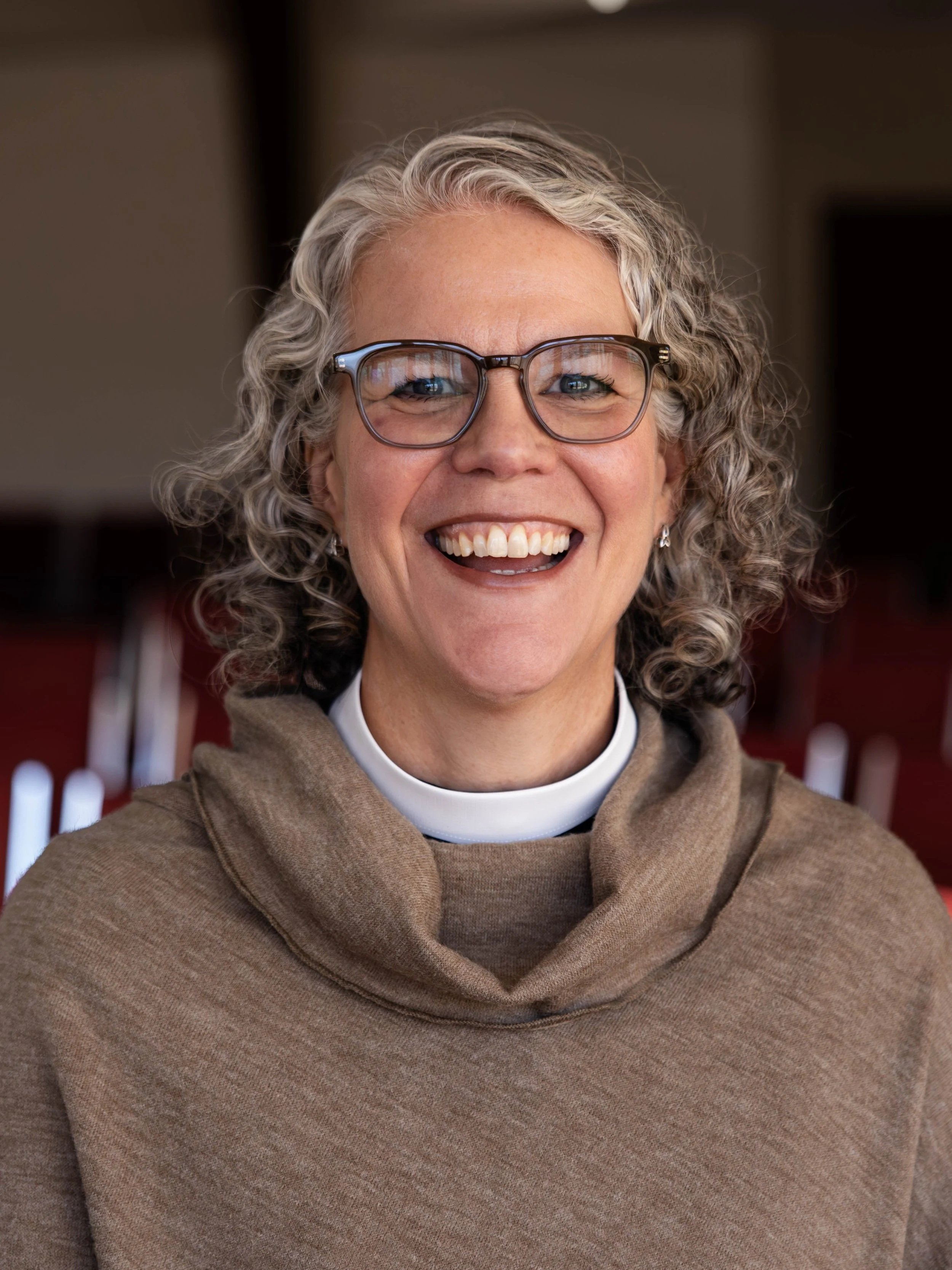 A woman with curly gray hair and glasses, smiling widely, wearing a brown cowl neck sweater and a white clerical collar.
