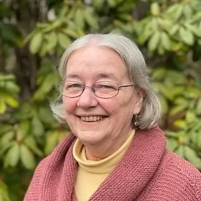 A smiling elderly woman with gray hair, glasses, wearing a pink cardigan and a yellow top, standing outdoors with green foliage in the background.