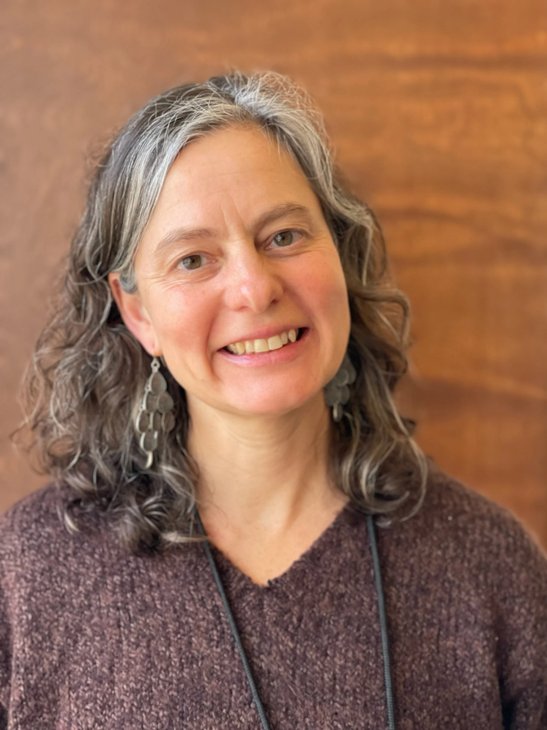 A woman with shoulder-length curly gray hair, wearing dangling earrings and a brown sweater, smiling at the camera in front of a wooden background.