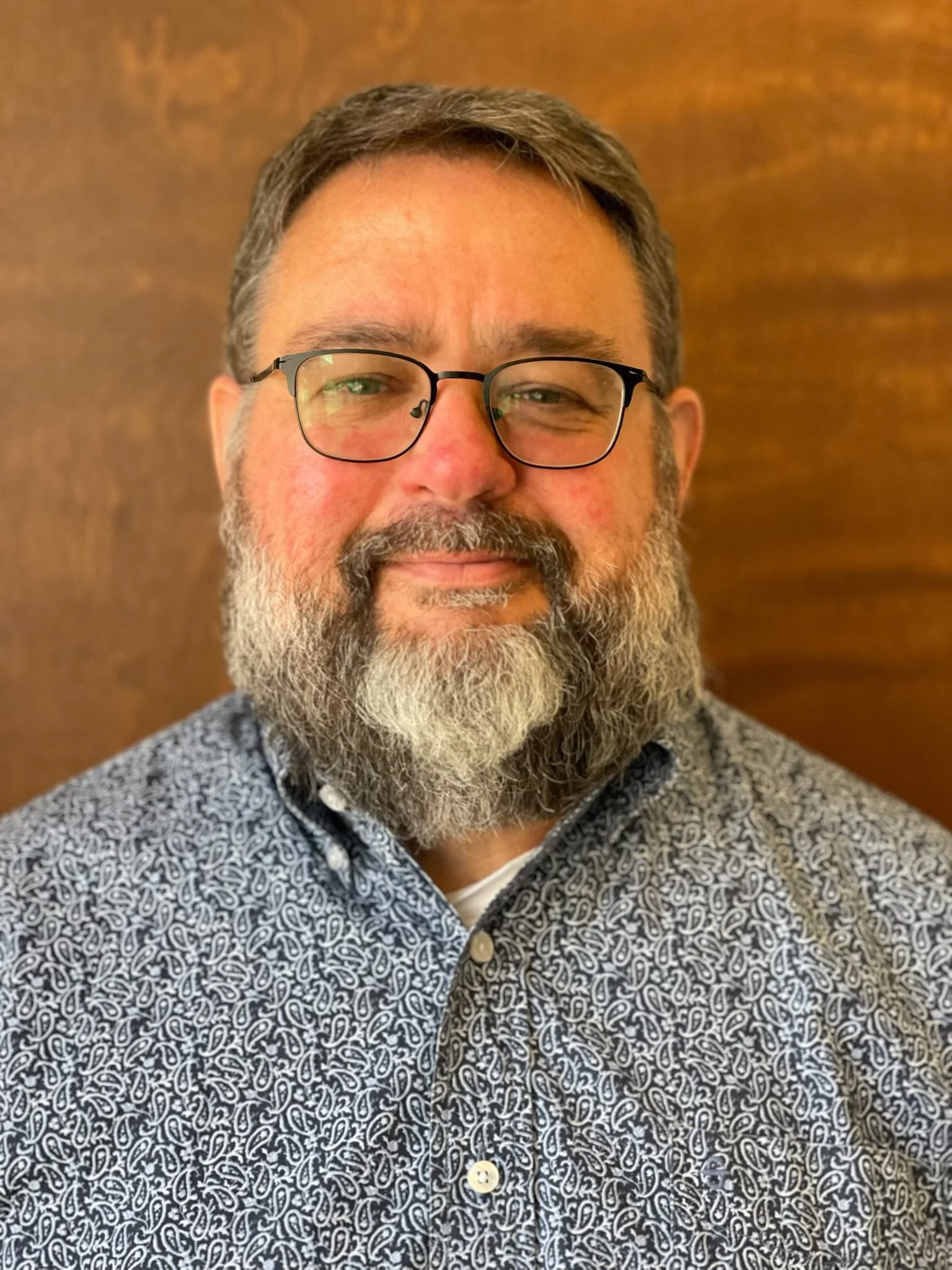 A middle-aged man with glasses, a full beard, and mustache, wearing a patterned button-up shirt, standing against a wooden background.