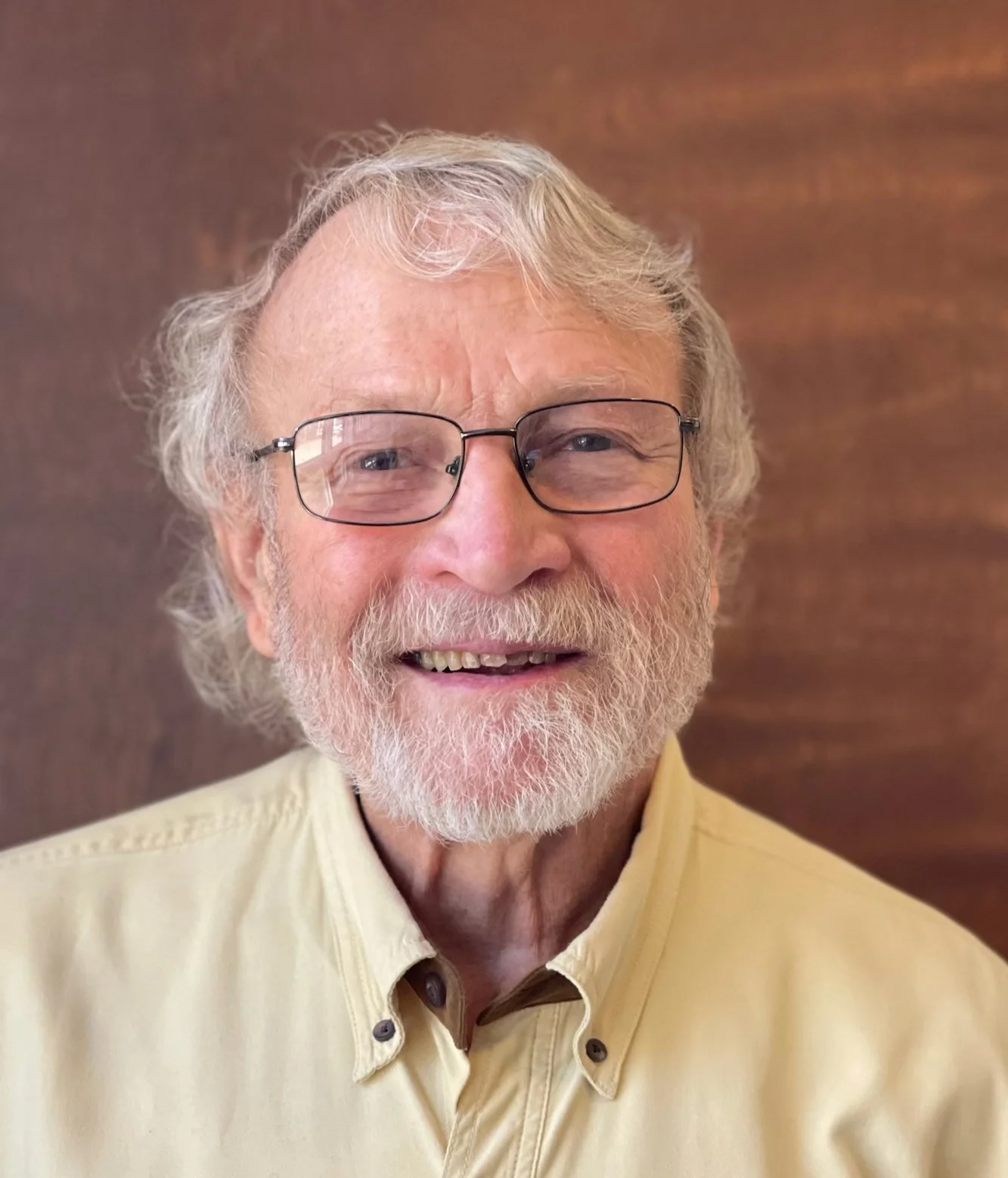 Close-up photo of smiling elderly man with glasses, white hair, and beard, wearing a light yellow collared shirt, in front of a wooden background.