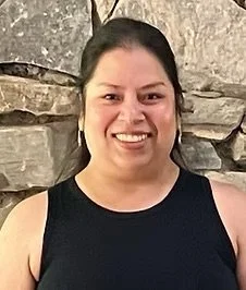 A woman smiling in front of a stone wall, wearing a sleeveless black top and gold hoop earrings.