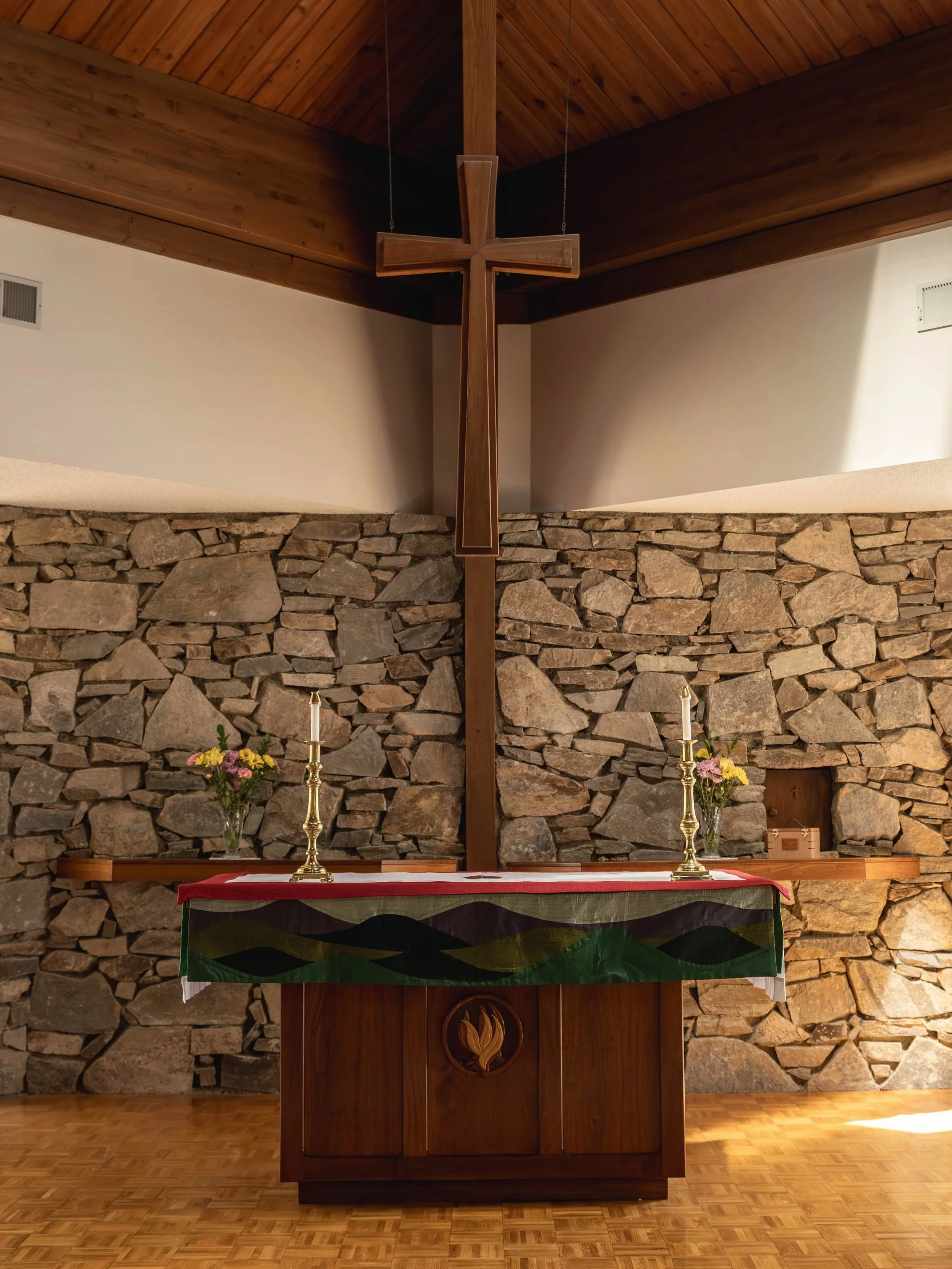 Christian altar with a large wooden cross, two altar candles, and flower arrangements in vases, set against a stone wall and wooden ceiling in a church interior.
