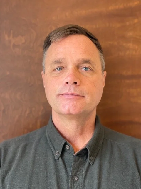 A man with short brown hair and blue eyes wearing a dark gray button-down shirt, standing in front of a wooden background.
