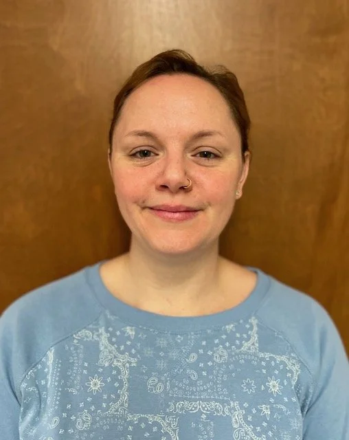 A woman with short brown hair, fair skin, a nose ring, and a subtle smile, wearing a light blue shirt with white patterns, standing in front of a wooden background.