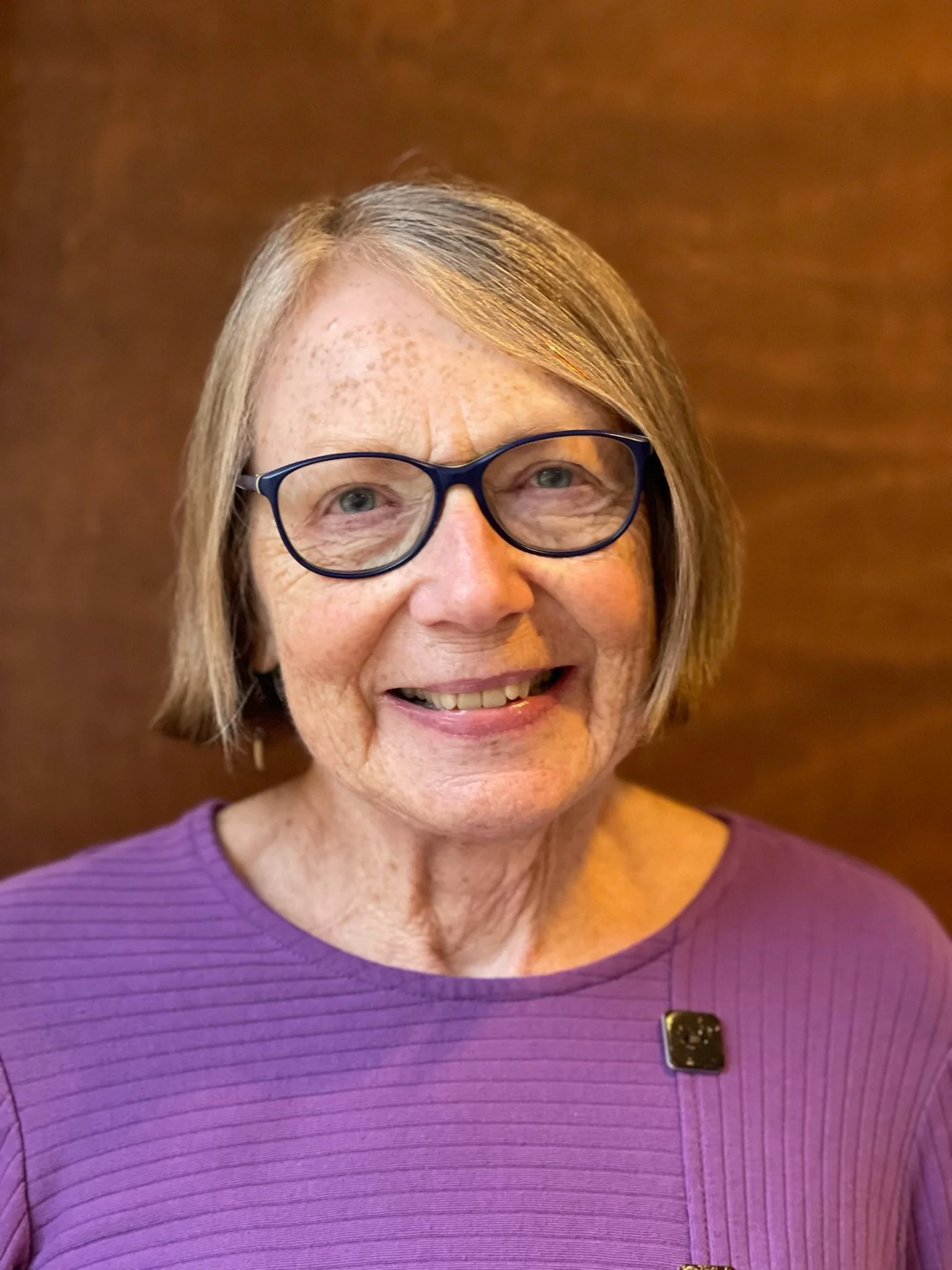 Smiling elderly woman with short gray hair, wearing glasses and a purple top, standing against a wooden background.