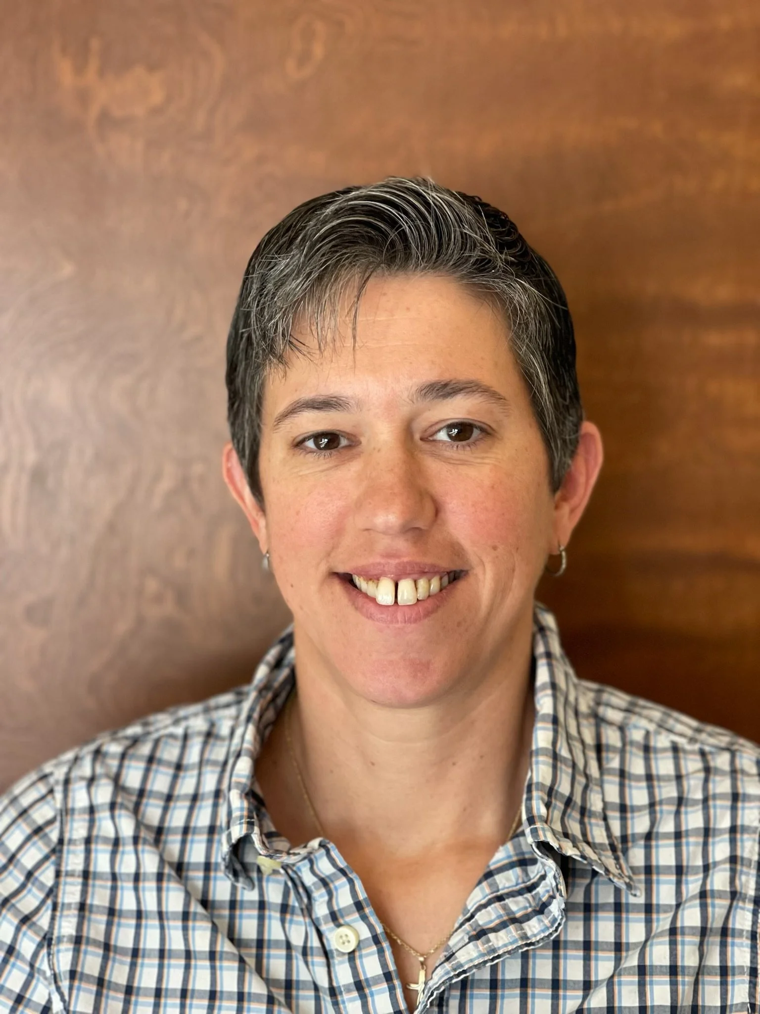A woman with short, dark hair and earrings, smiling, wearing a plaid shirt, standing against a wooden background.