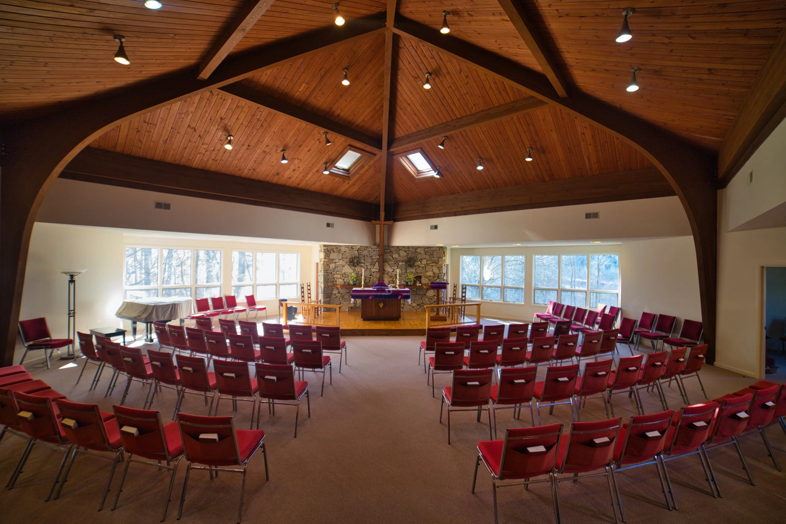 An indoor chapel or meeting space with rows of red chairs facing an altar with a stone wall backdrop. The ceiling is wooden with several skylights and hanging lights. Large windows on the side let in natural light, and there are trees visible outside.