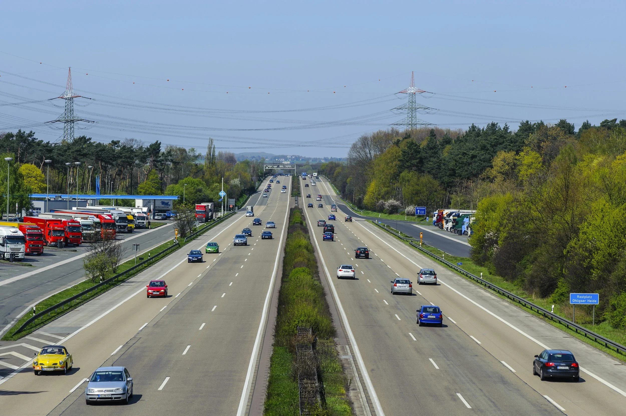 A highway with multiple lanes, some cars and trucks traveling on it, surrounded by green trees and electrical towers under a clear blue sky.
