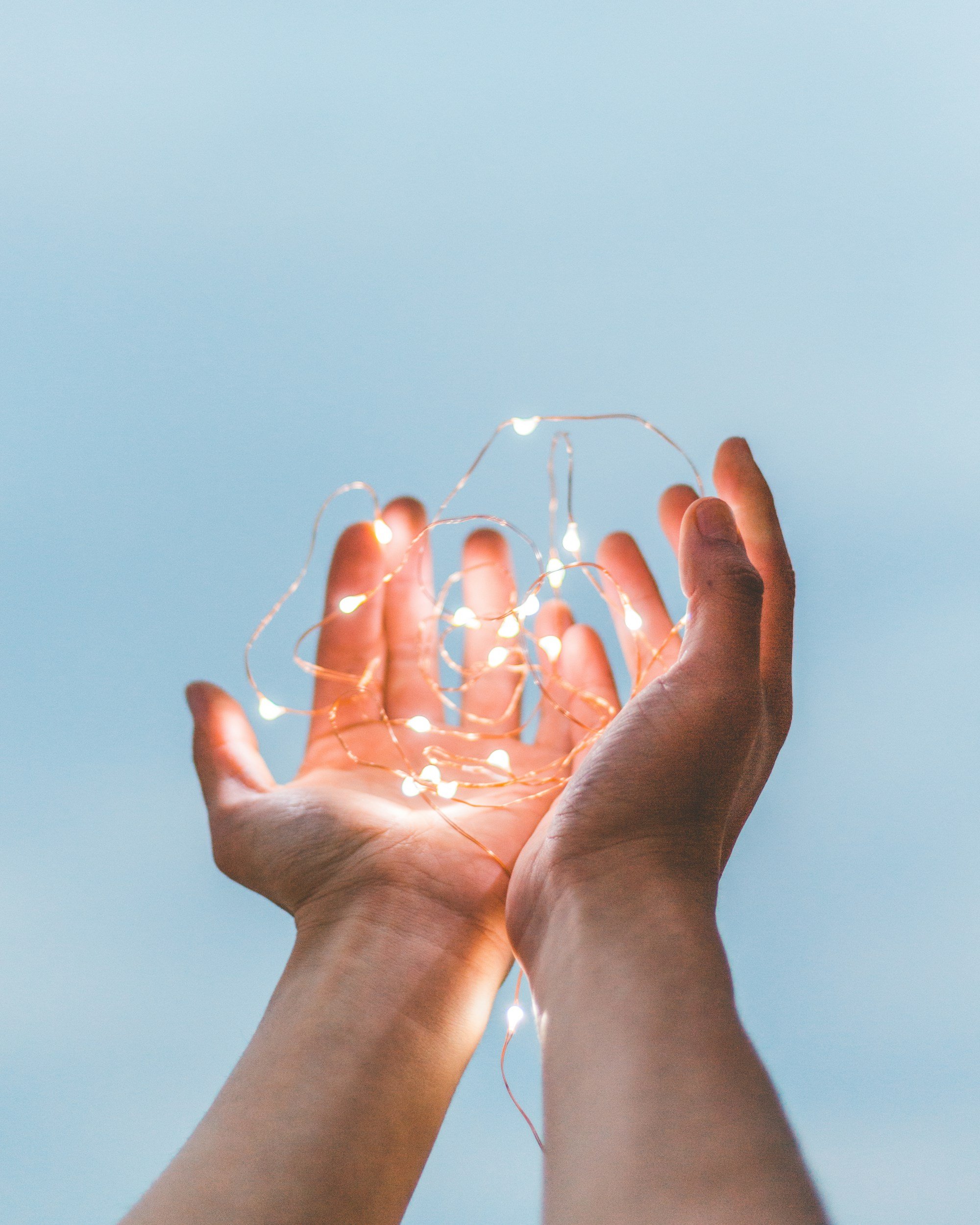 A person's hands holding a tangled string of small, glowing fairy lights against a light blue background.