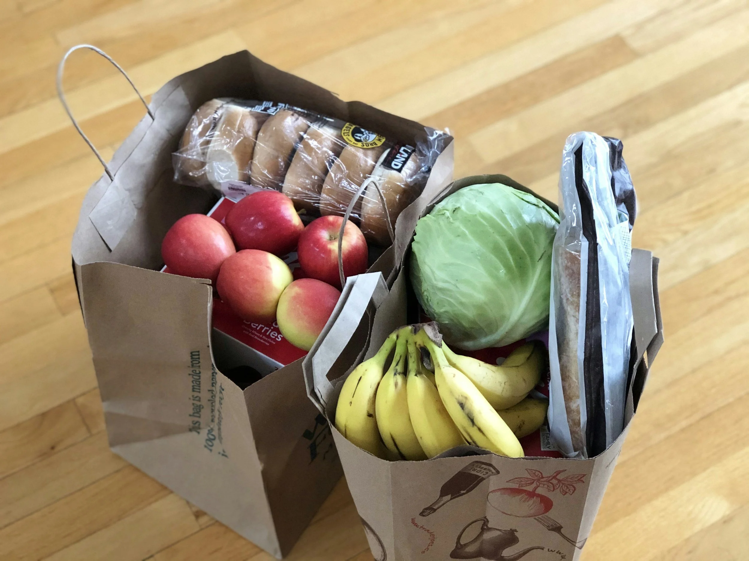 Two paper grocery bags filled with fruits and vegetables, including apples, bananas, a head of cabbage, and a bag of bread rolls, on a wooden floor.