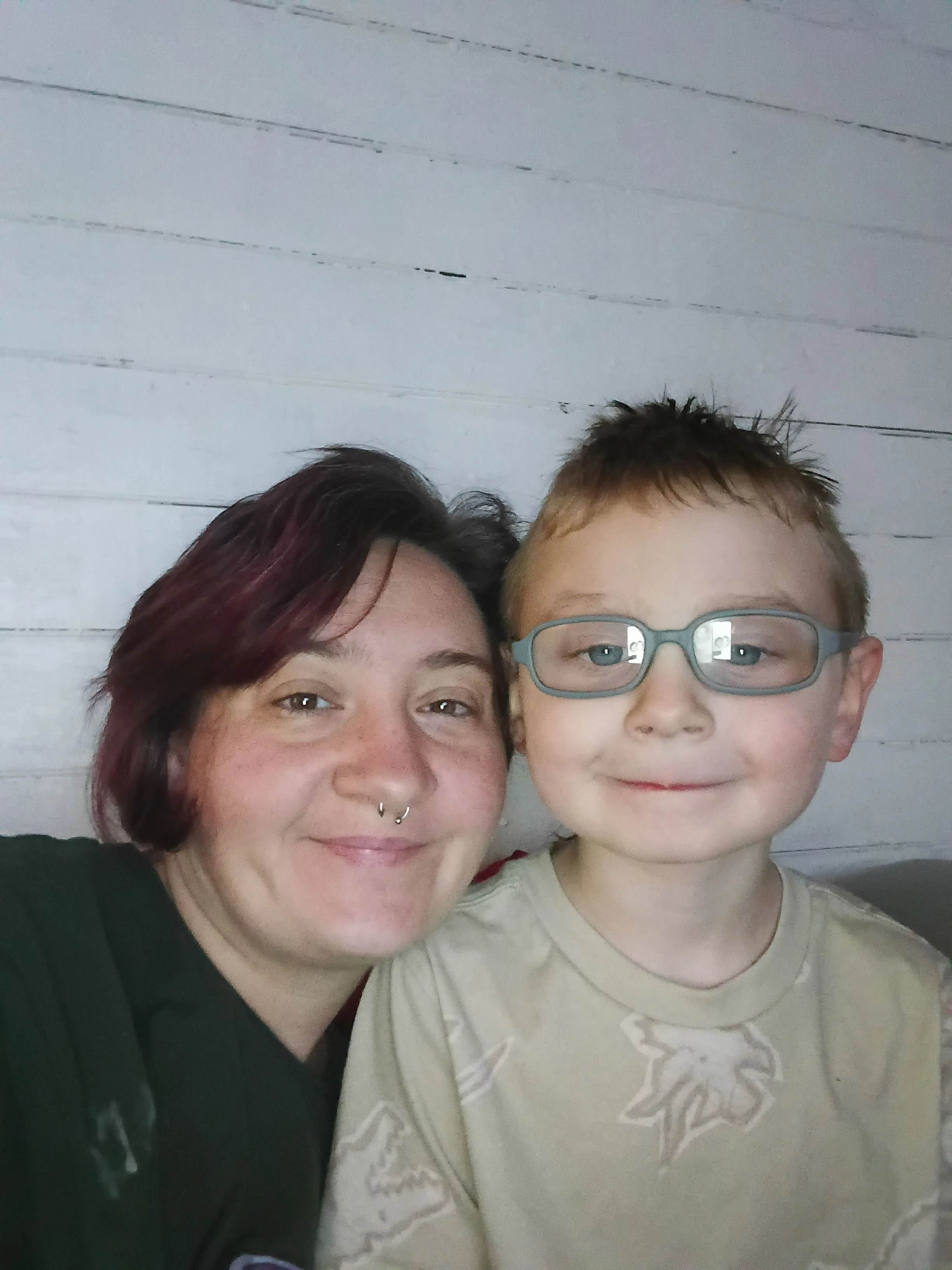 A woman with short, dark hair and a septum piercing smiling next to a young boy with glasses and short, light brown hair, both smiling at the camera, with a white wooden wall in the background.