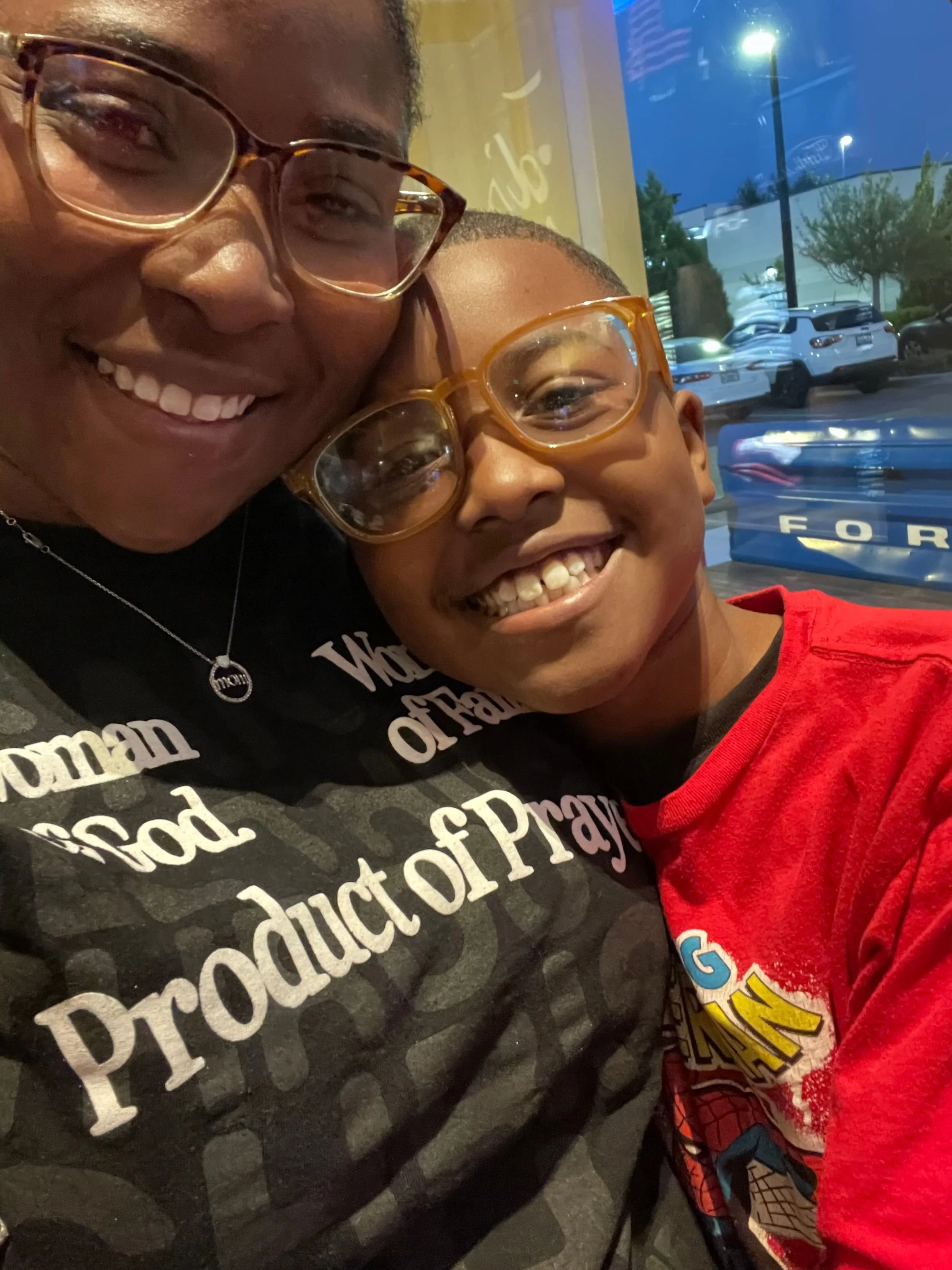 A woman and a young boy wearing glasses are smiling and posing together for a selfie inside a restaurant. The woman is wearing a black shirt with white text, and the boy is wearing a red shirt with a graphic design. They are close to each other, showing happiness.