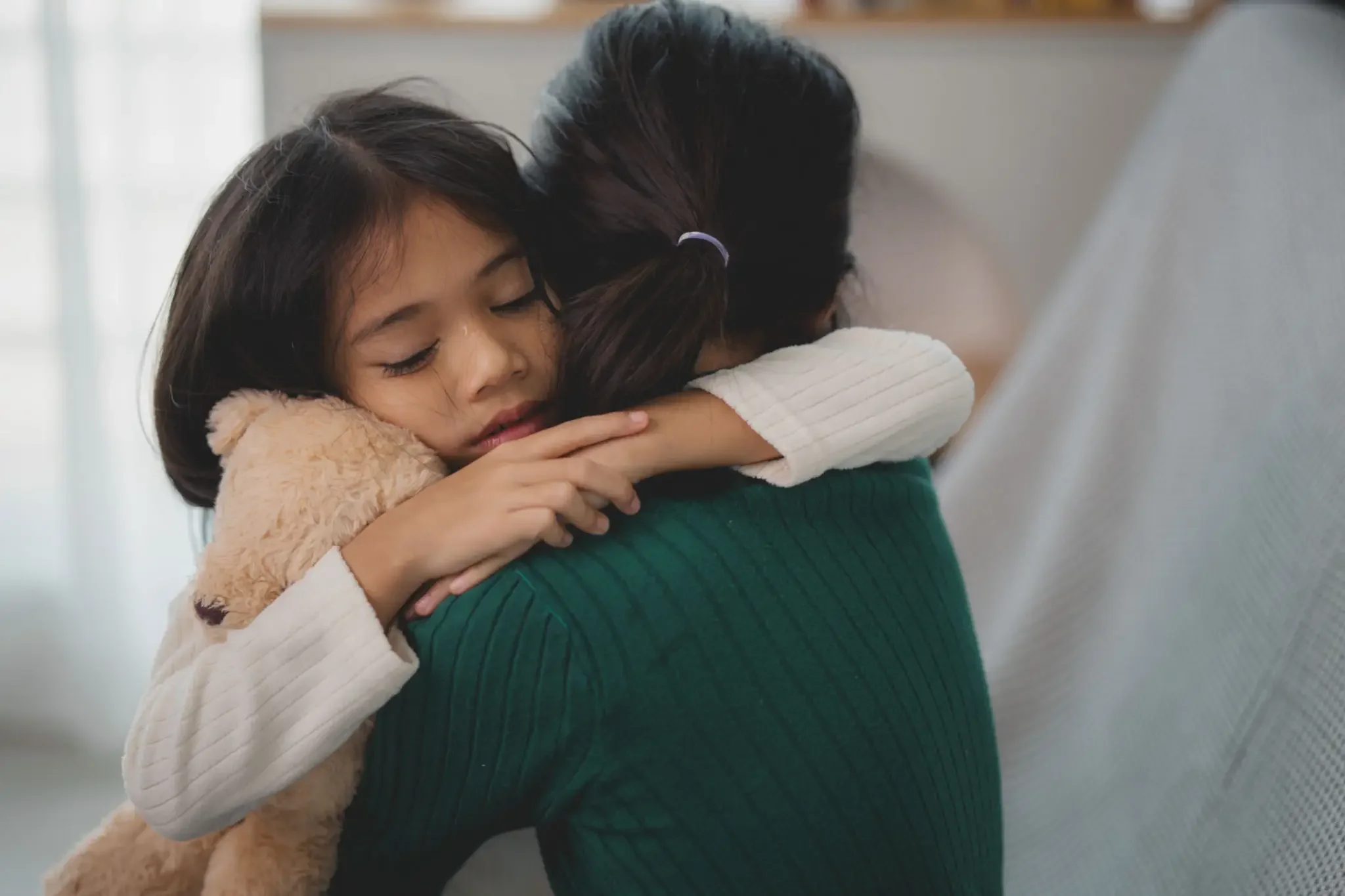 A young girl with dark hair hugging an adult woman, their eyes closed in an emotional embrace.