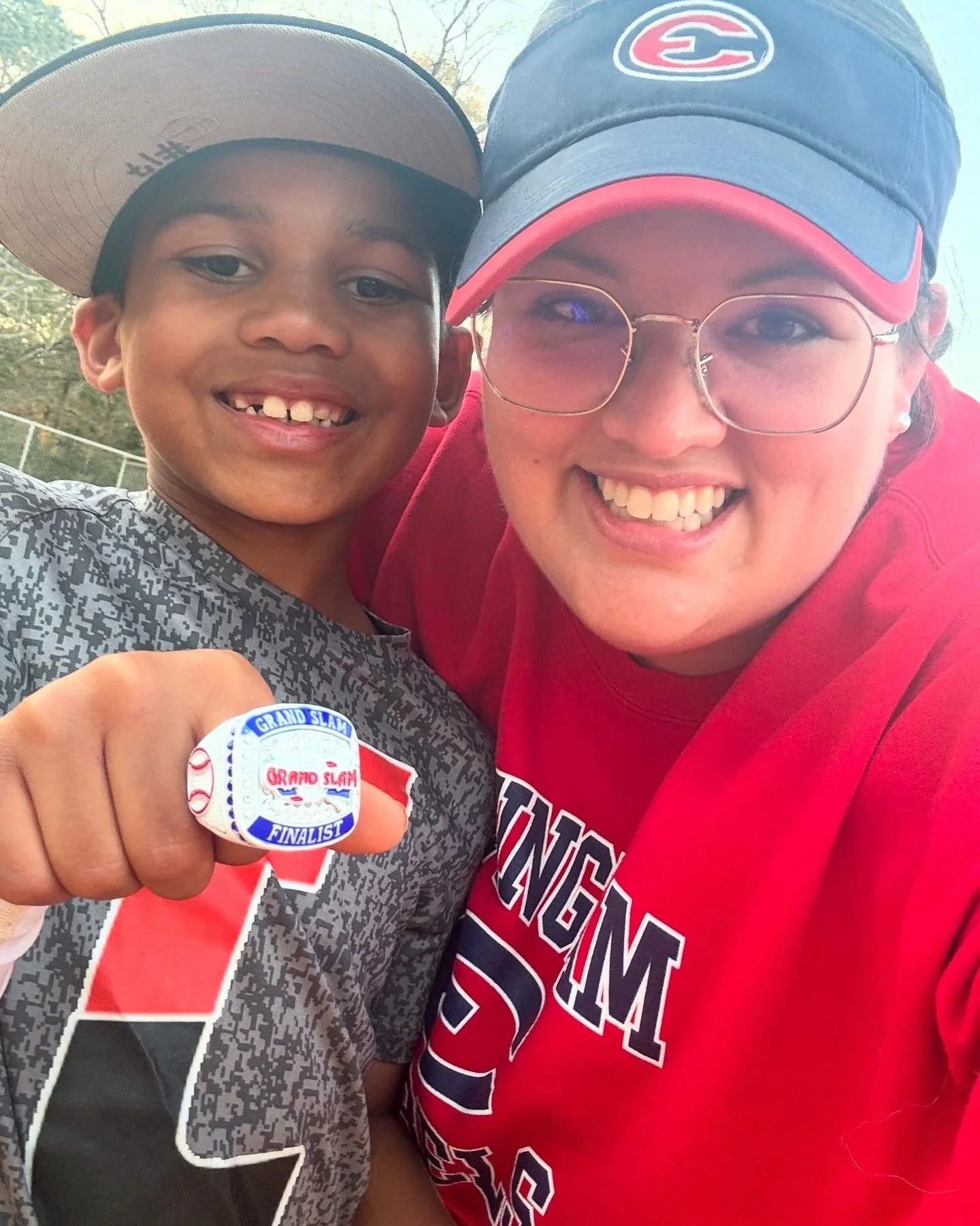 A young boy wearing a gray and black sports jersey and a tan cap holding a pin with 'Grand Slam Finalist' on it, smiling next to a woman in a red shirt and blue cap with glasses, both smiling.