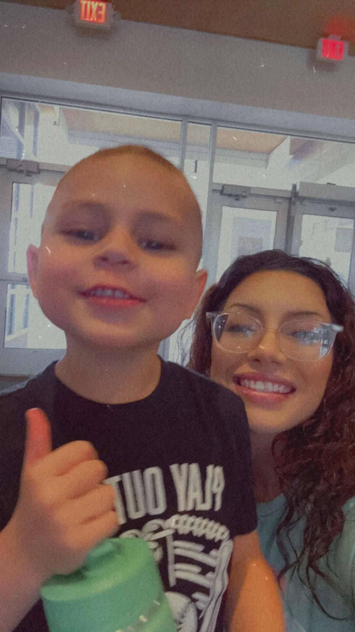 A young boy smiling and giving a thumbs-up, with a woman wearing glasses smiling in the background, inside a building with glass doors and exit signs.