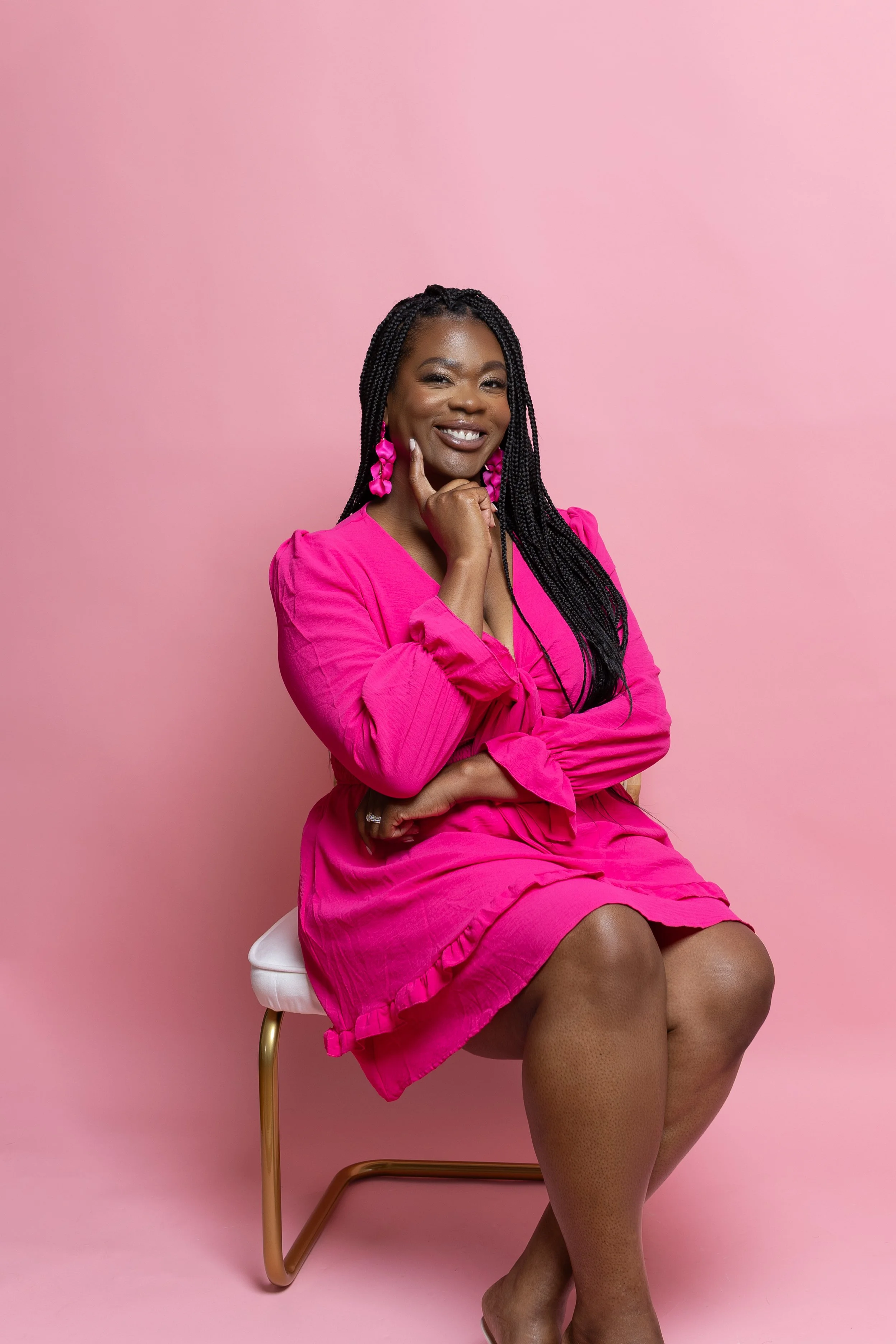 A woman with long braided hair wearing a vibrant pink dress and matching earrings, sitting on a white chair with gold legs against a pink background, smiling and posing with her hand near her chin.
